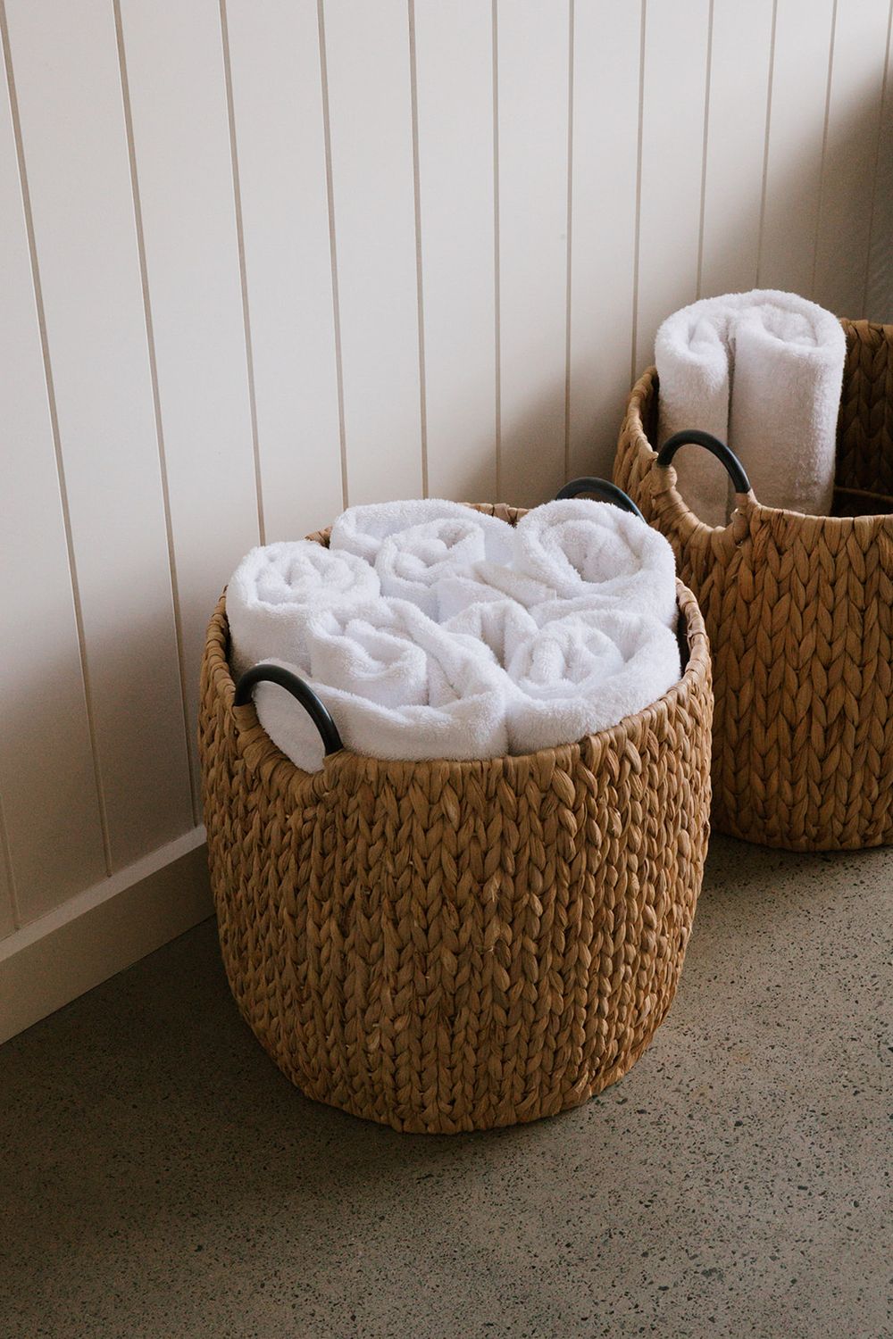 Woven seagrass baskets with black handles filled with rolled white towels, placed on a concrete floor against a white paneled wall.