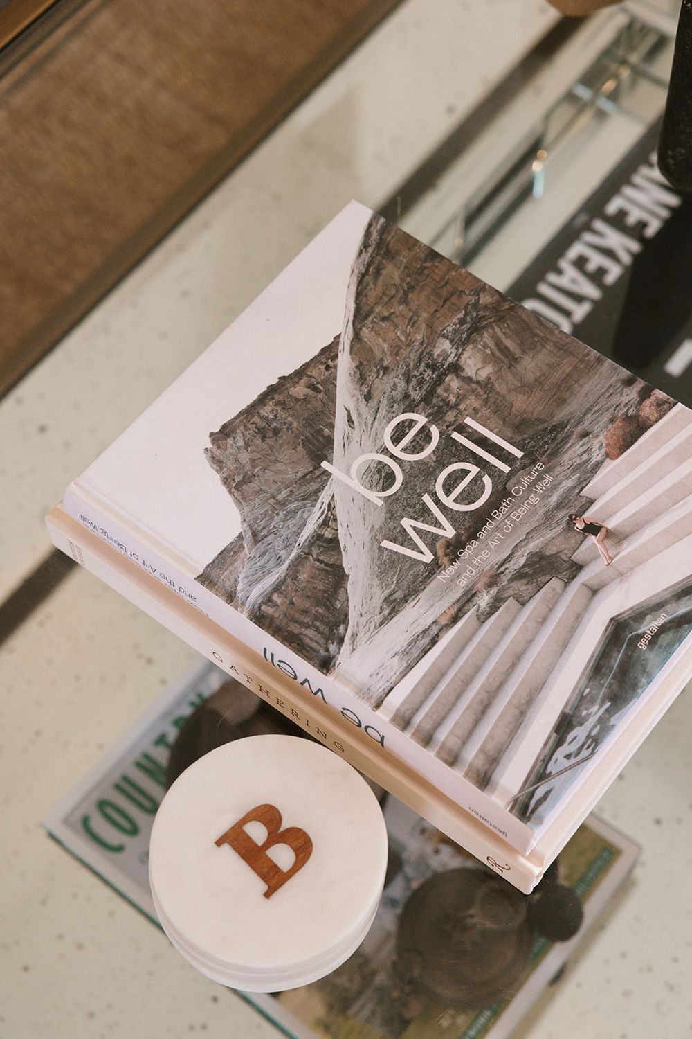 Stack of wellness books including 'Be Well' on a glass coffee table with a marble coaster featuring letter B.