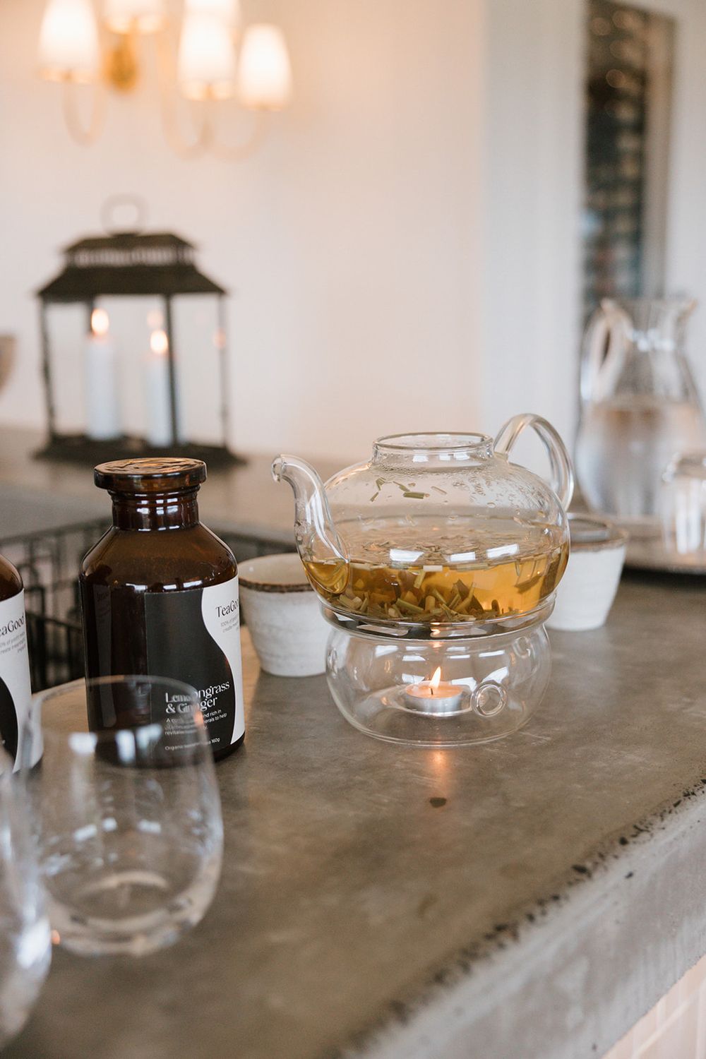 Glass teapot with herbal tea on a candle warmer beside amber bottles and glassware on a concrete surface.