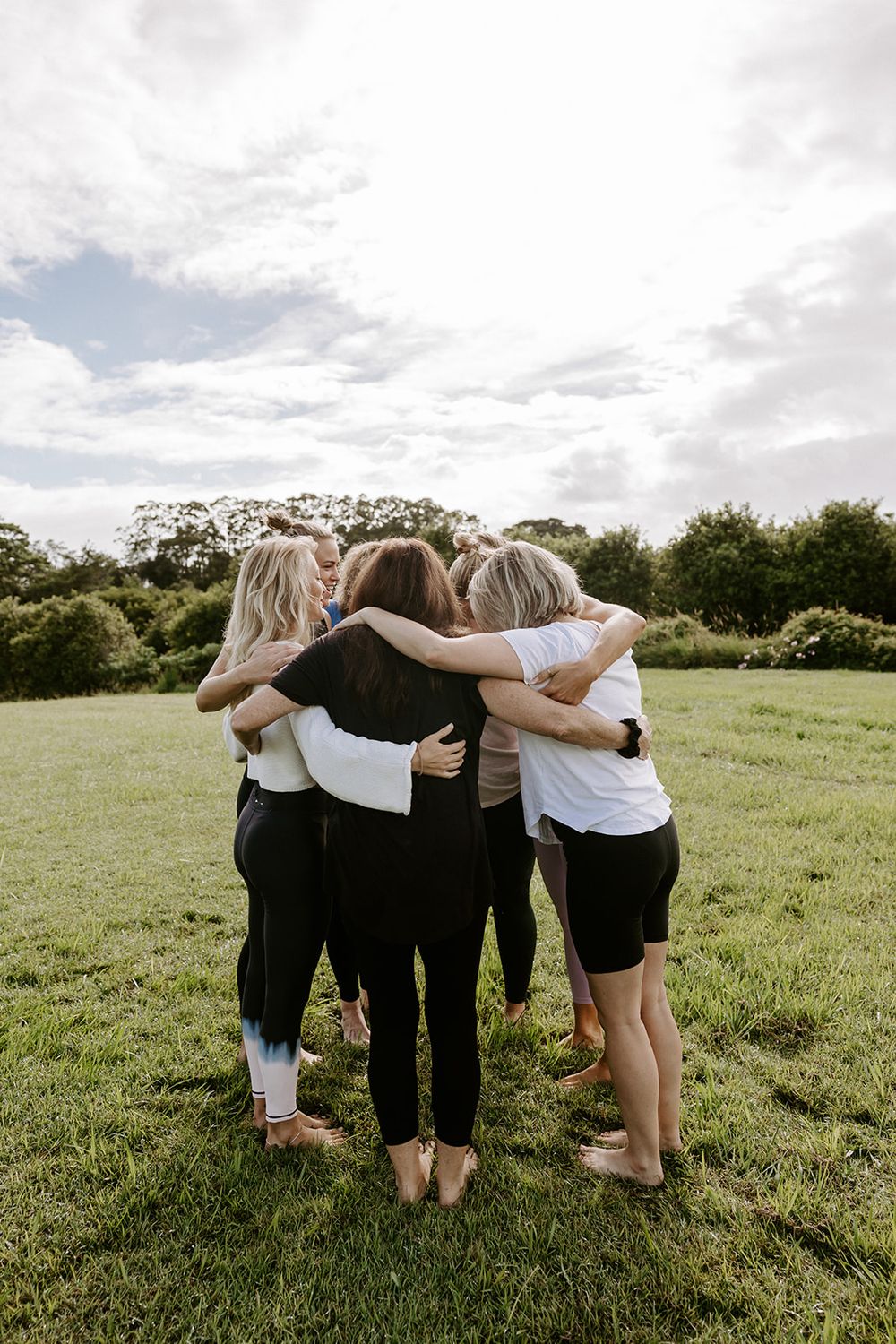 Group of women hugging in a circle on green grass field with trees and cloudy sky in background.