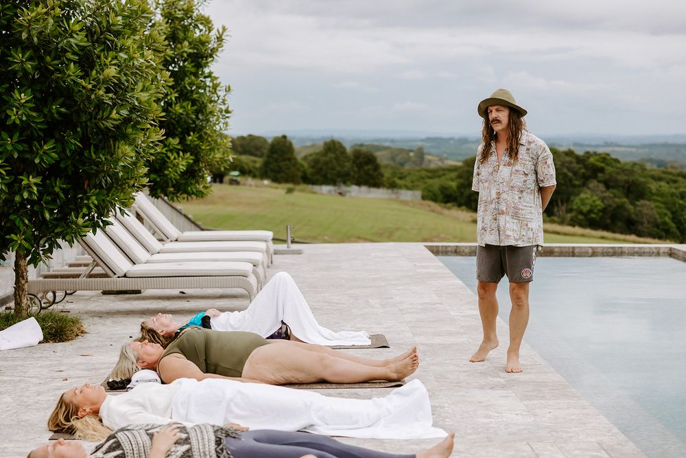 People relaxing by an outdoor pool on lounge chairs while a man with long hair stands nearby on a sunny day.