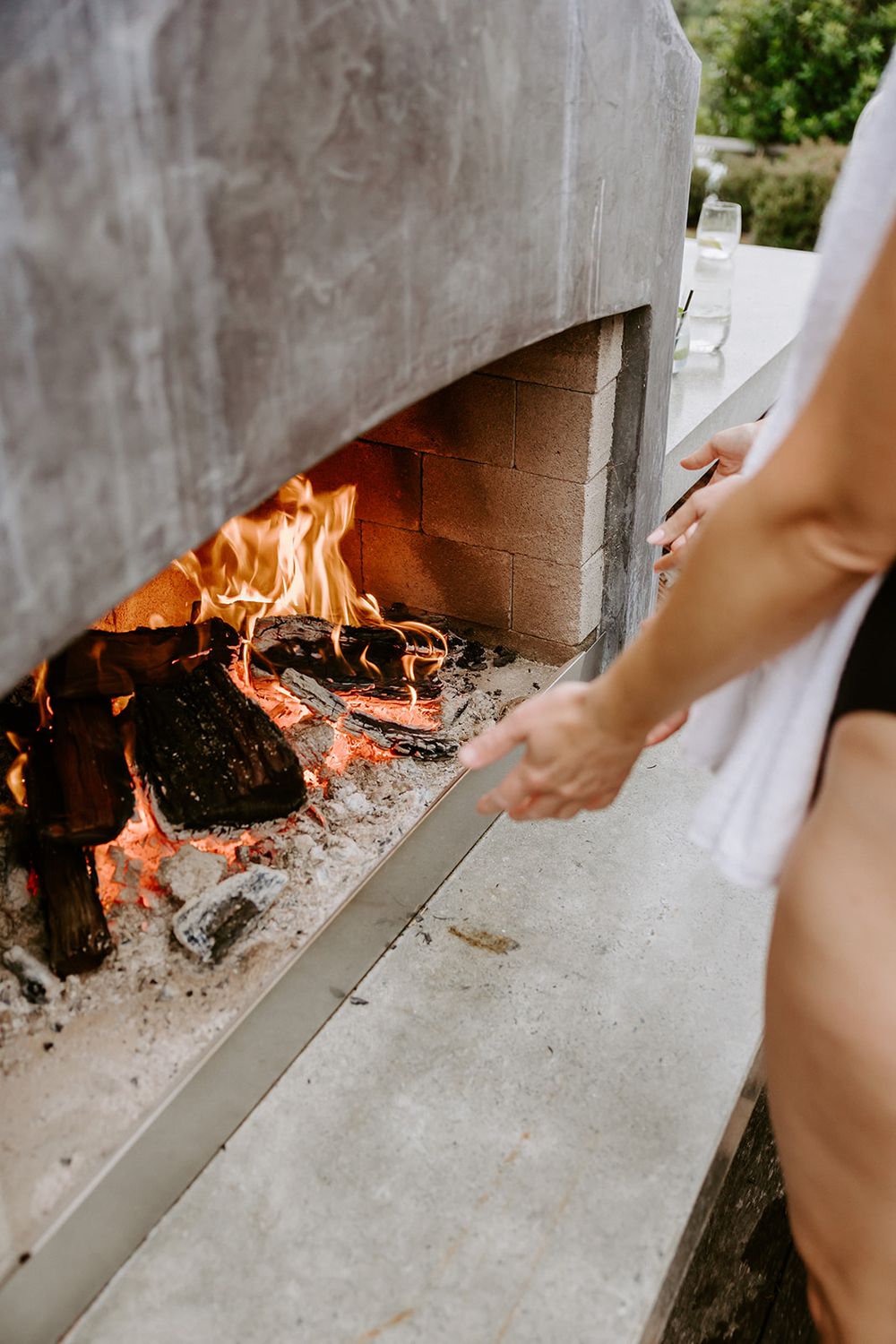Person tending to a wood-fired outdoor pizza oven with bright orange flames burning inside a concrete structure.