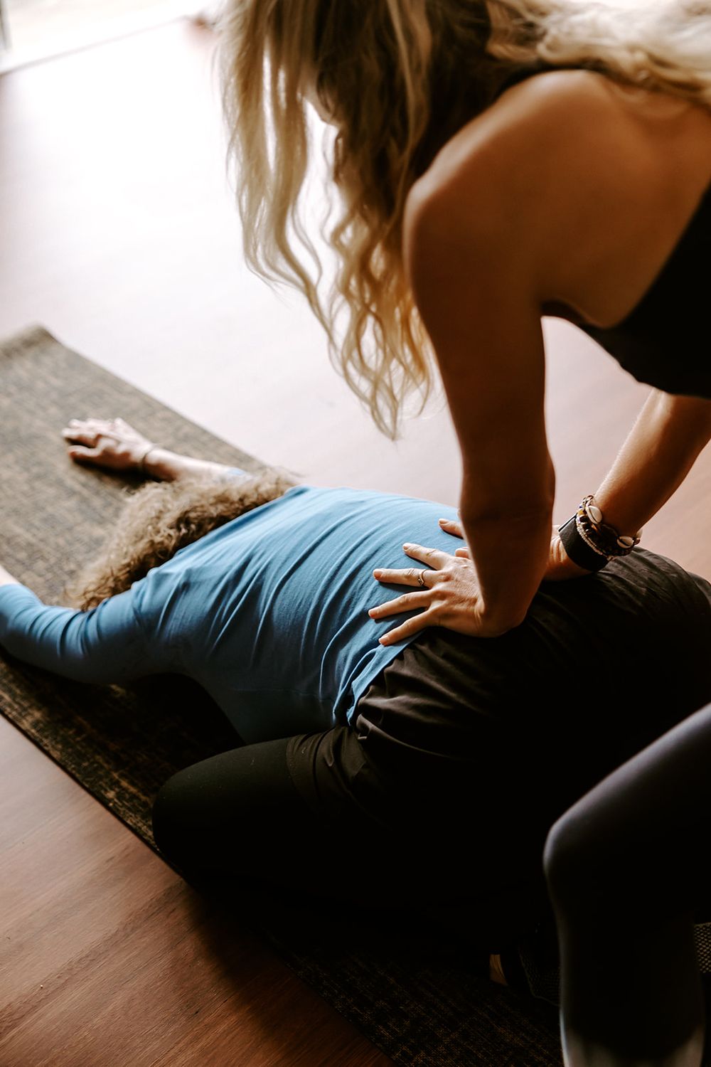 A yoga instructor assists a student lying on a mat, applying hands-on adjustment to their lower back during a session.