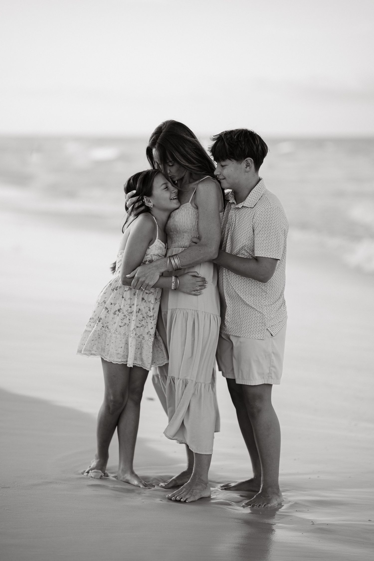 Black and white photo of a mother and two children sharing a tender embrace on a sandy beach shore.