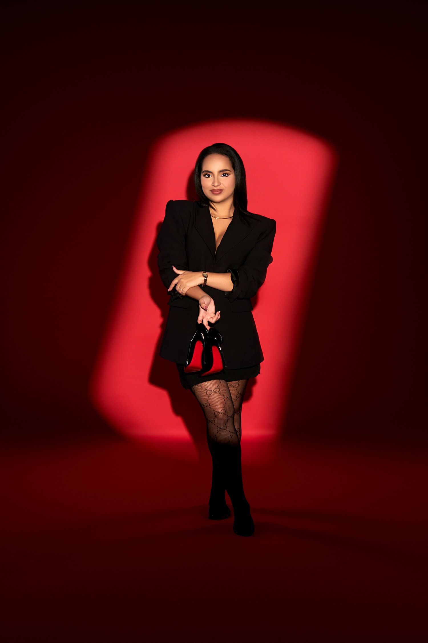 A woman in black blazer dress and fishnet stockings poses confidently against a dramatic red spotlight background.