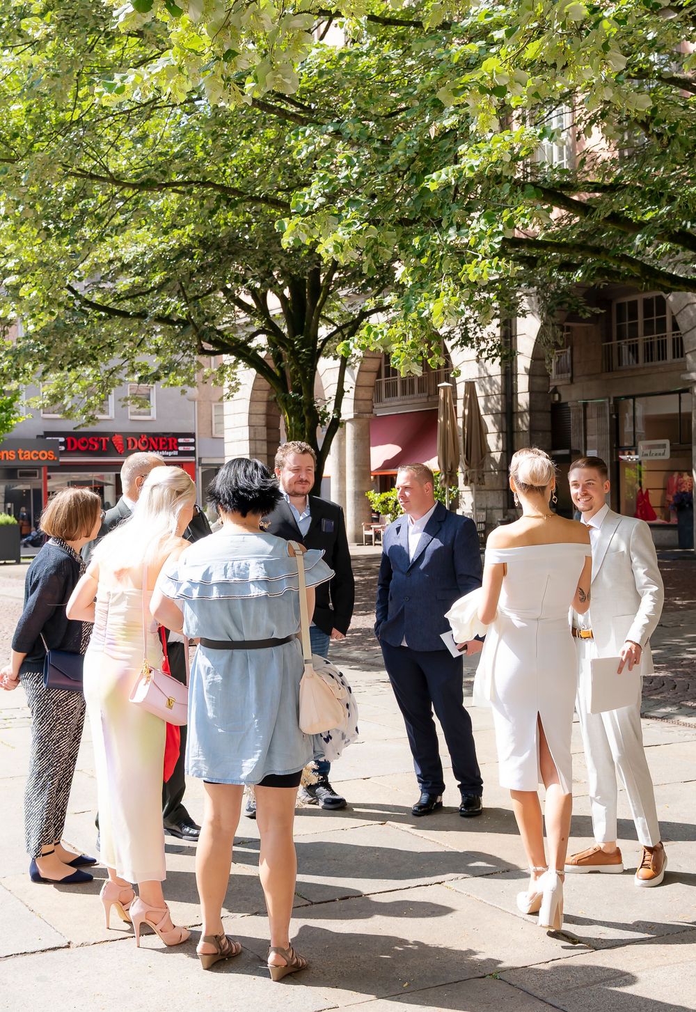 Wedding guests mingling outdoors under green trees on a sunny day.