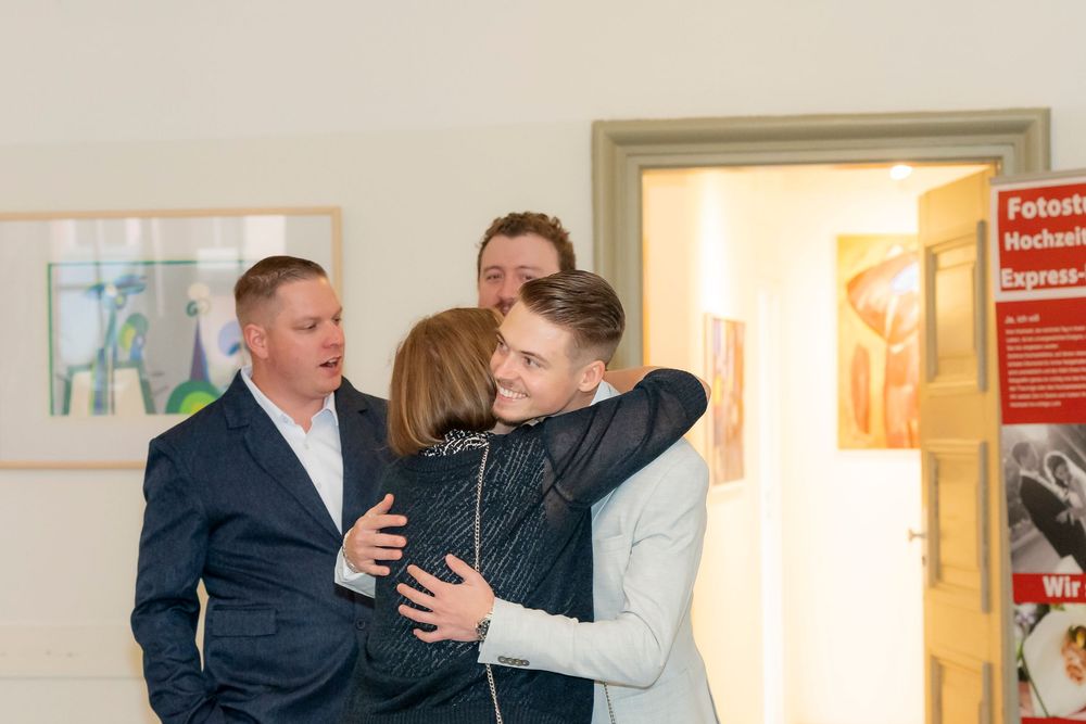 People in formal attire interacting in a hallway with light colored walls.