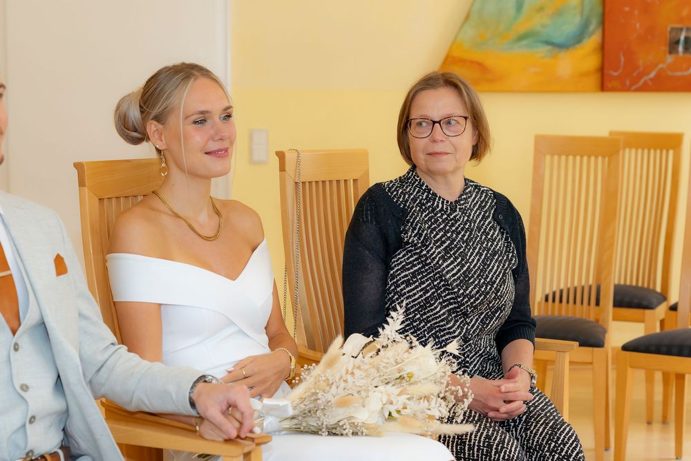 Wedding attendees seated in wooden chairs with yellow walls and colorful artwork in the background.