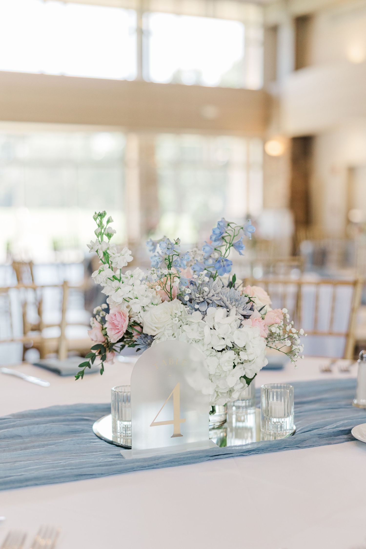 Elegant wedding reception table with blue runner, white and pink floral centerpiece, candles, and gold chairs in bright venue.