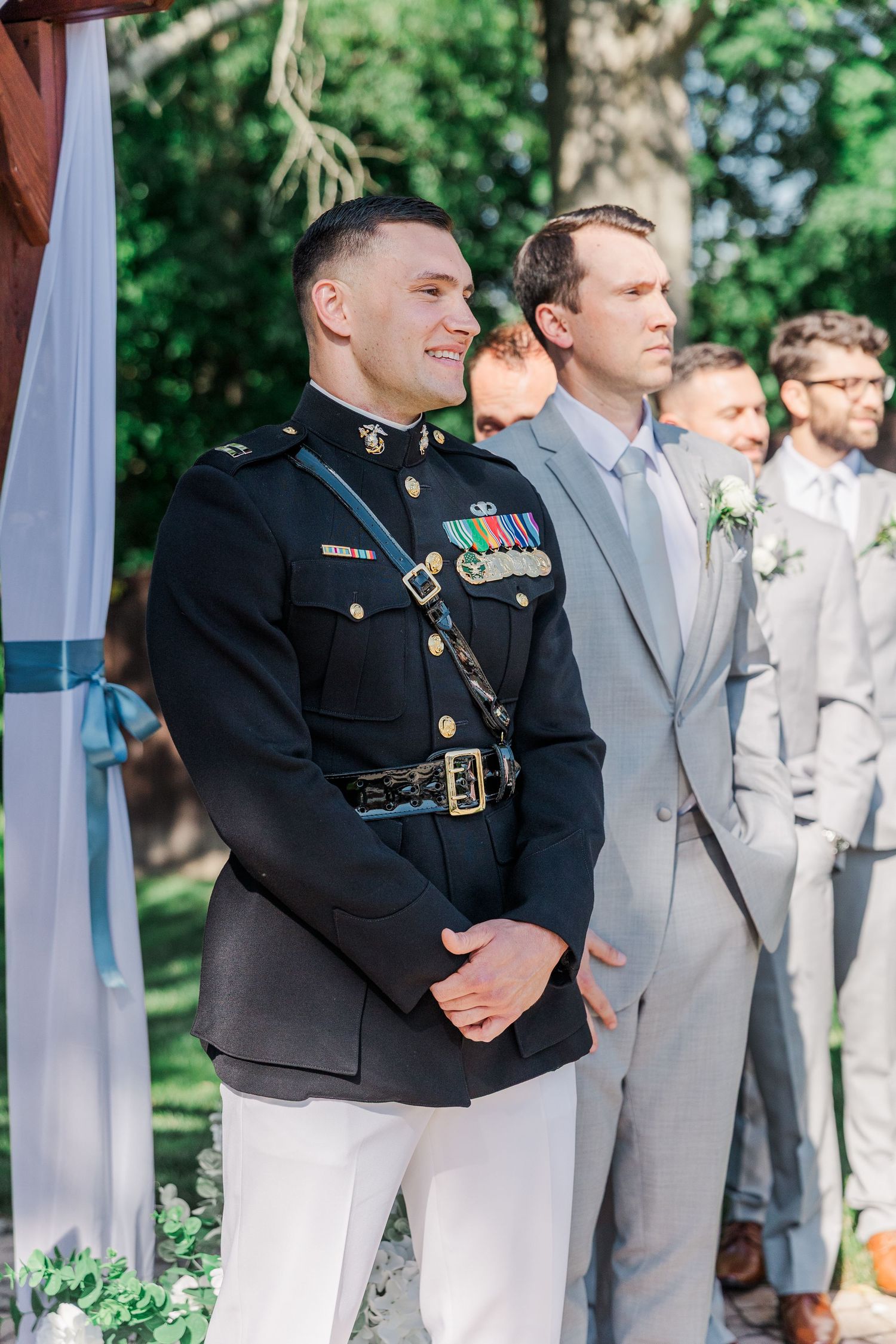 Military groom in dress uniform smiles at altar beside groomsmen in gray suits at outdoor wedding ceremony.