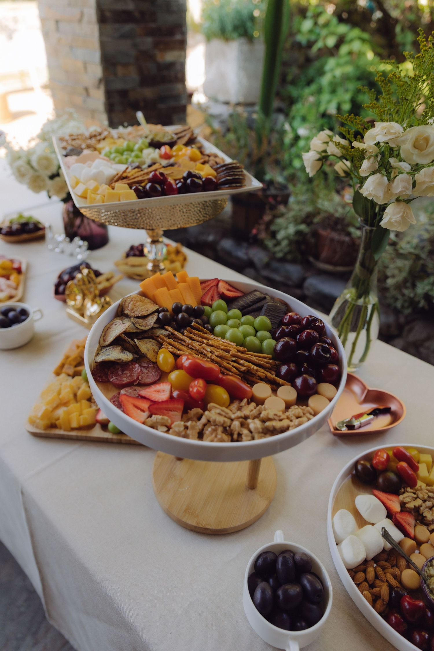 Wedding reception grazing table featuring tiered platters with grapes, cherries, crackers, nuts and assorted charcuterie outdoors.