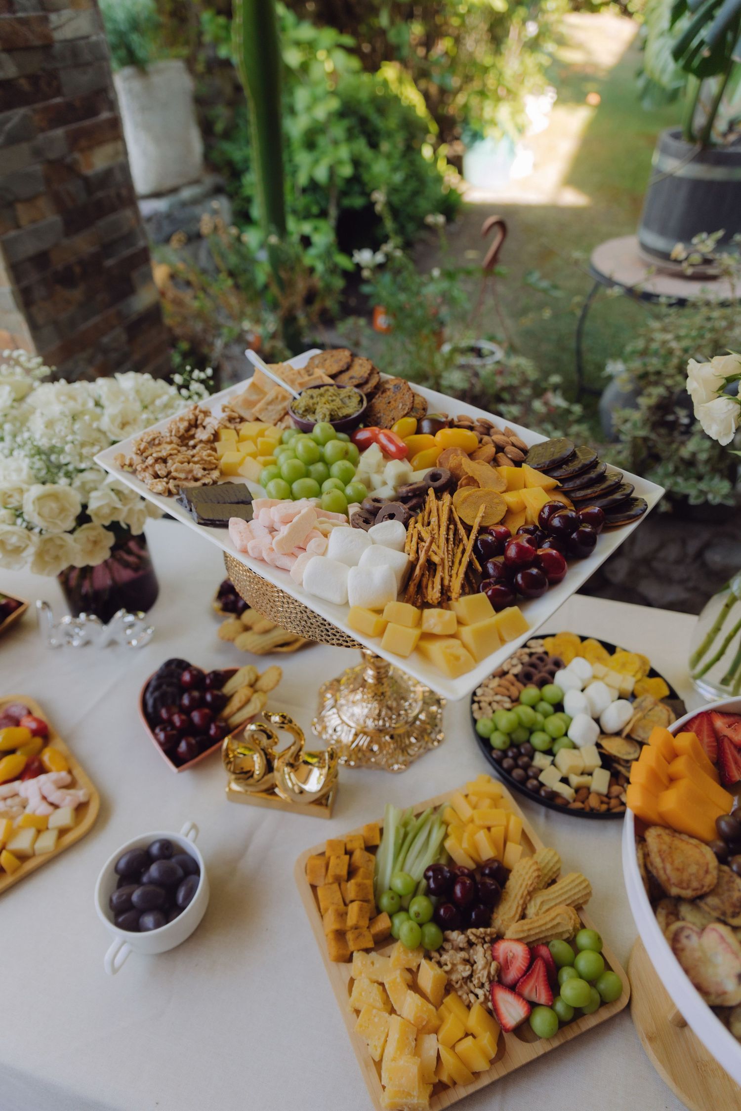 Outdoor wedding reception food table with charcuterie boards filled with grapes, cheeses, fruits and crackers surrounded by flowers.