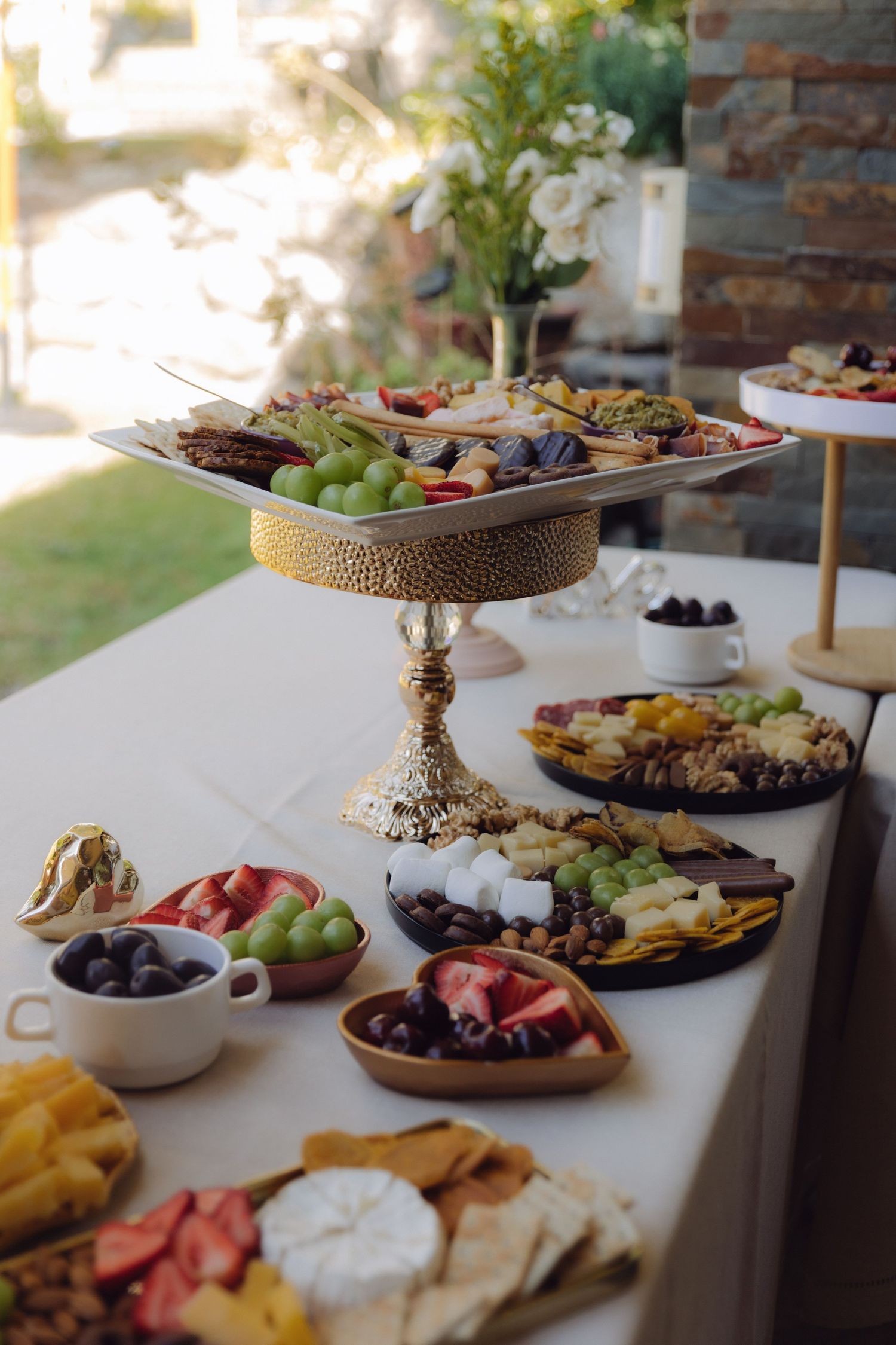 Elegant outdoor wedding grazing table with tiered gold stand displaying fruits, cheeses, crackers and charcuterie arrangements.