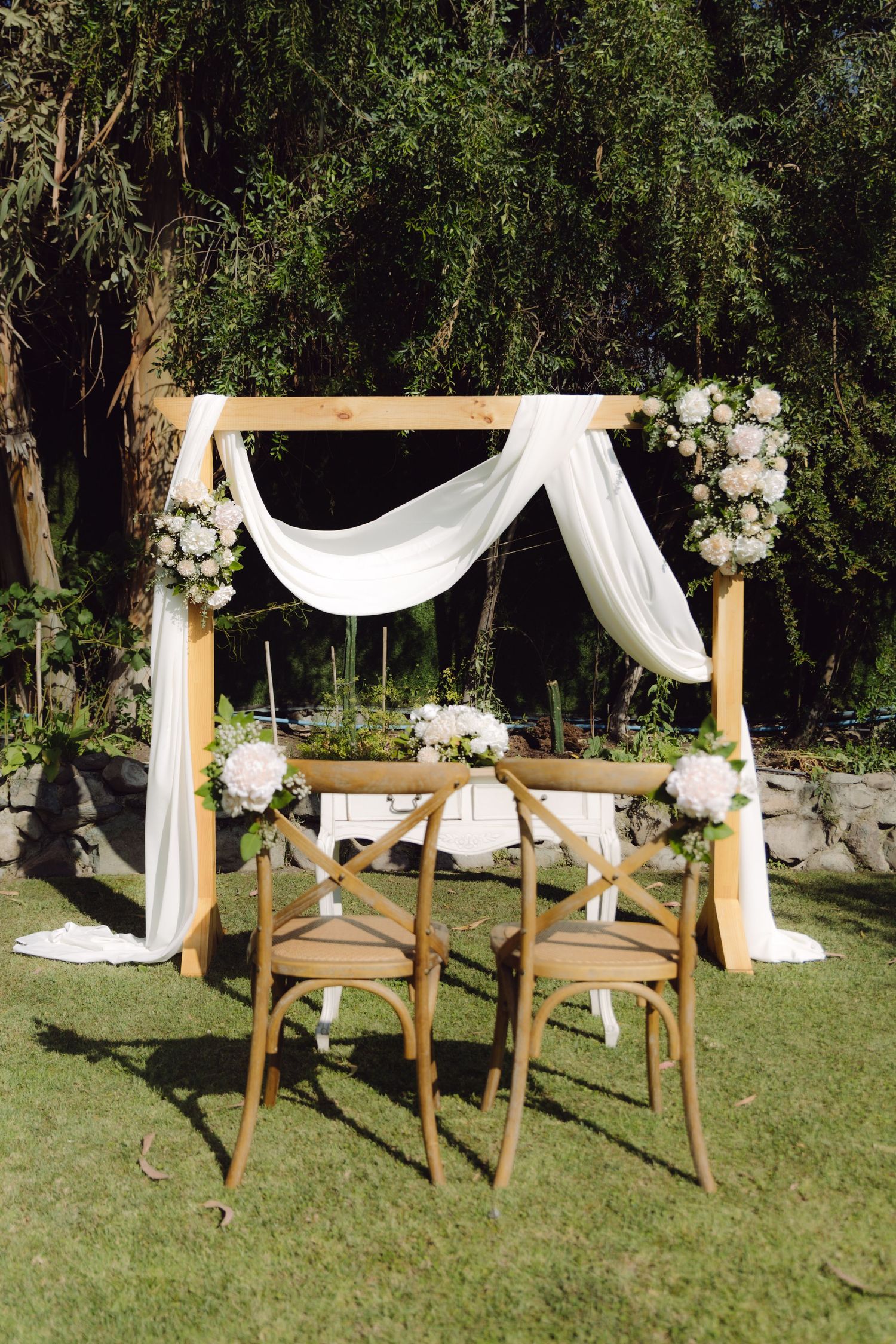 Outdoor wedding ceremony arch decorated with white fabric draping, floral arrangements and two wooden cross-back chairs on green lawn.