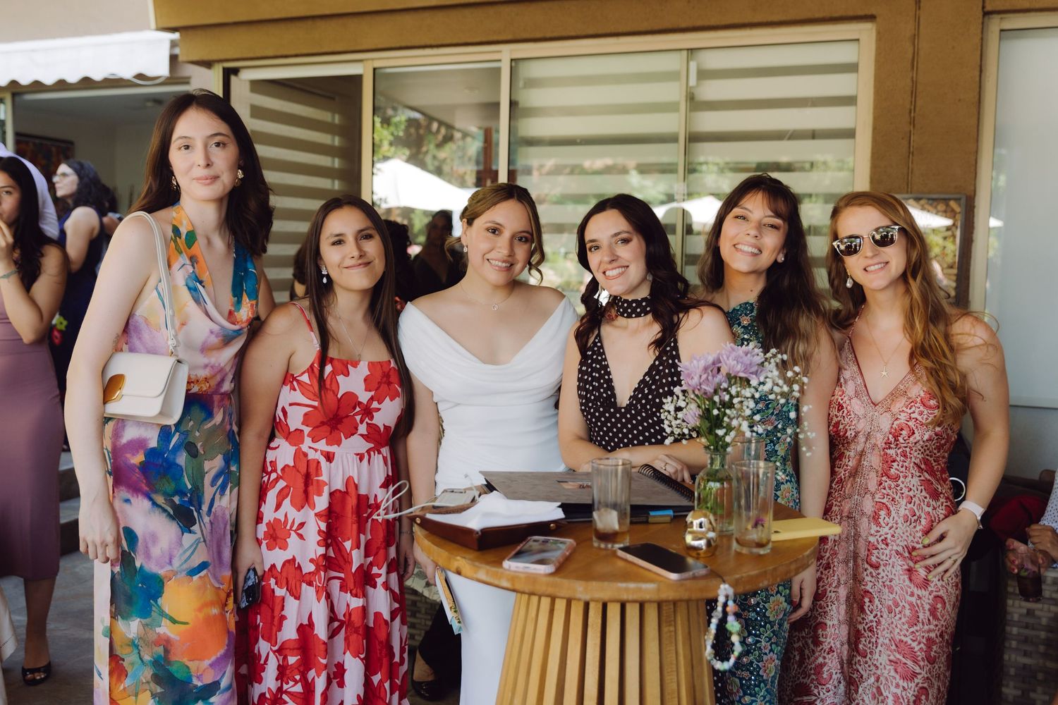 Bride in white dress posing with five female wedding guests in colorful floral dresses at outdoor reception.