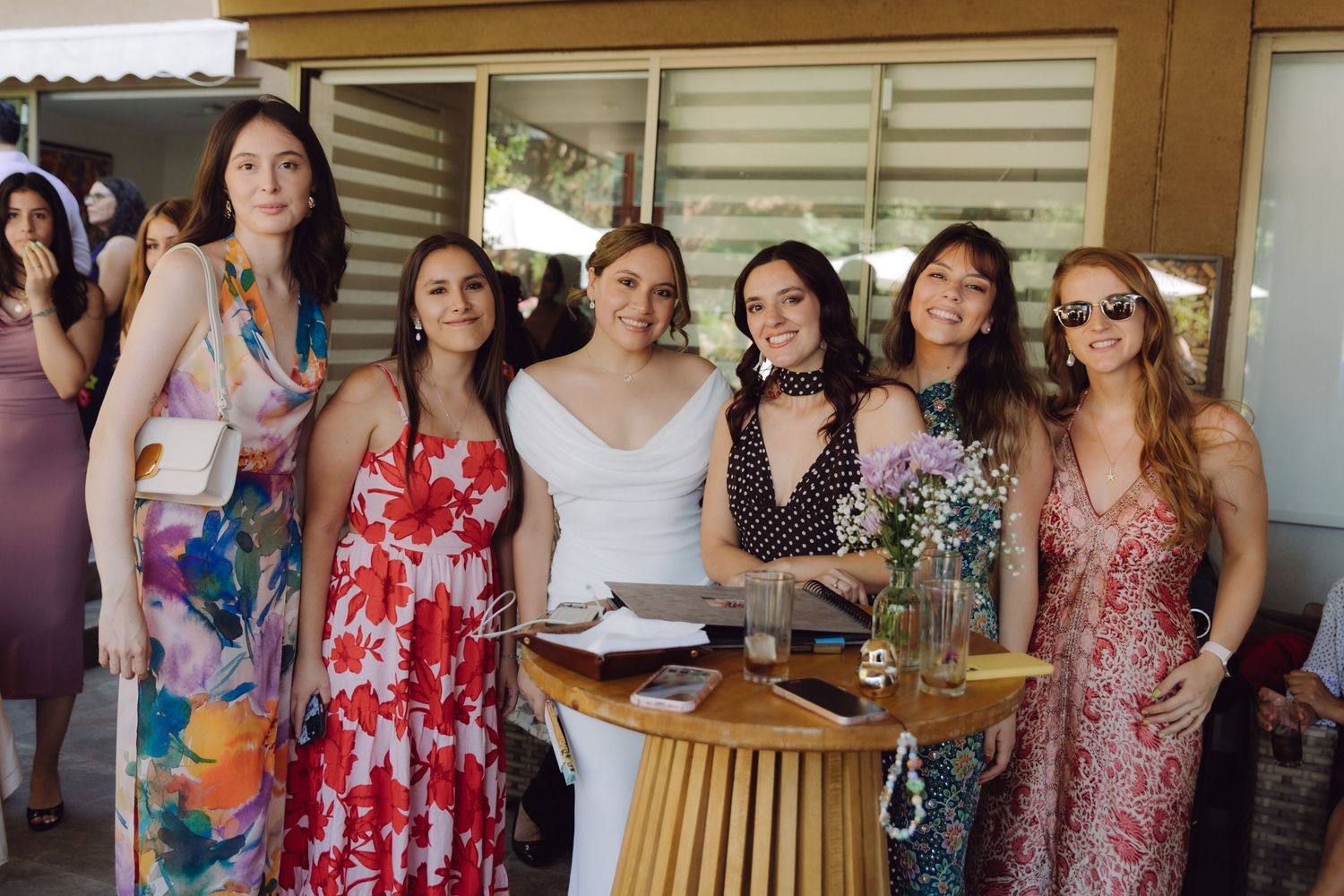 Bride in white dress smiling with five female guests wearing colorful summer dresses at outdoor wedding reception.
