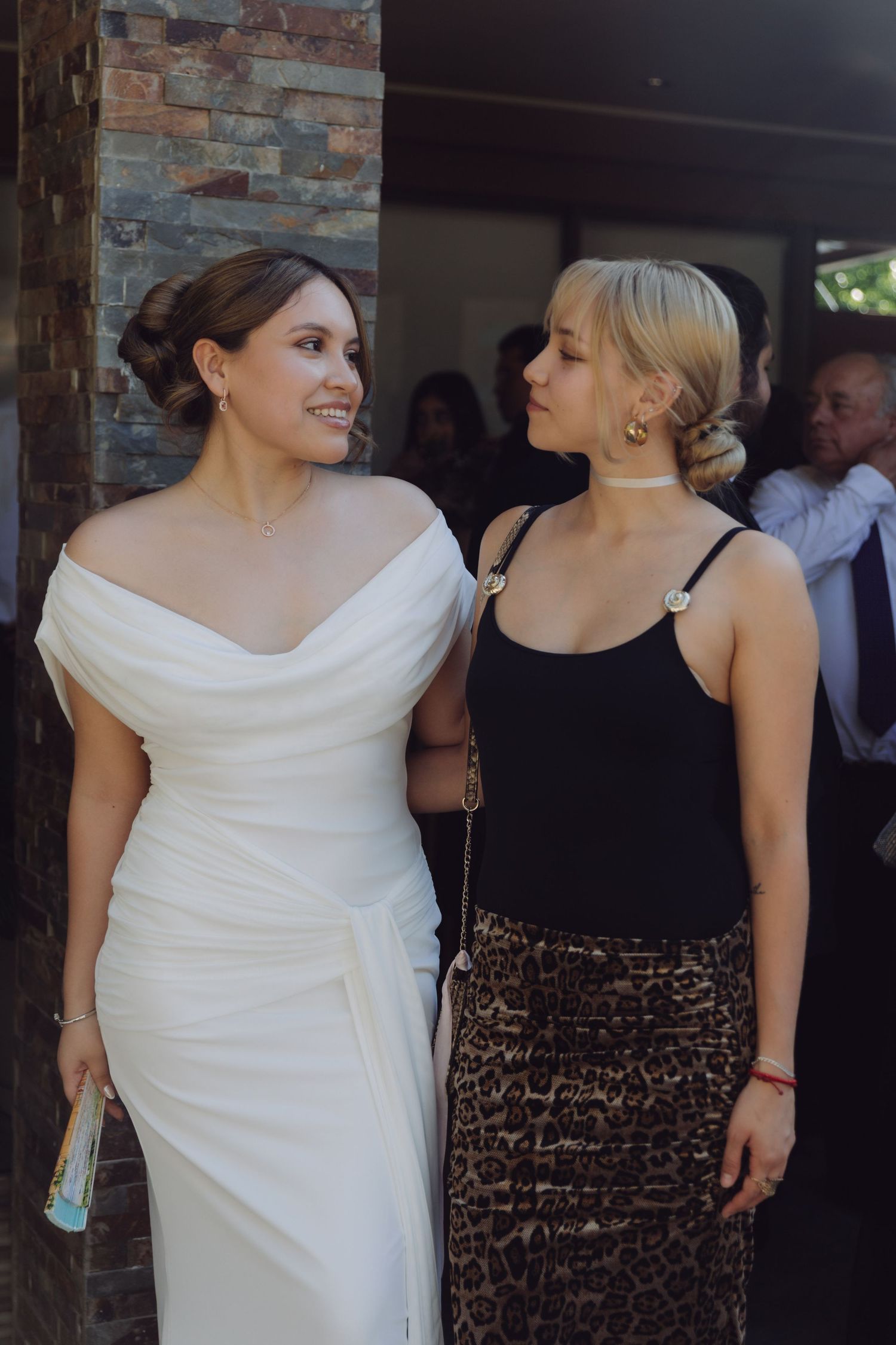 Two women at a wedding, one in a white bridal gown and one in a black top with leopard print skirt.
