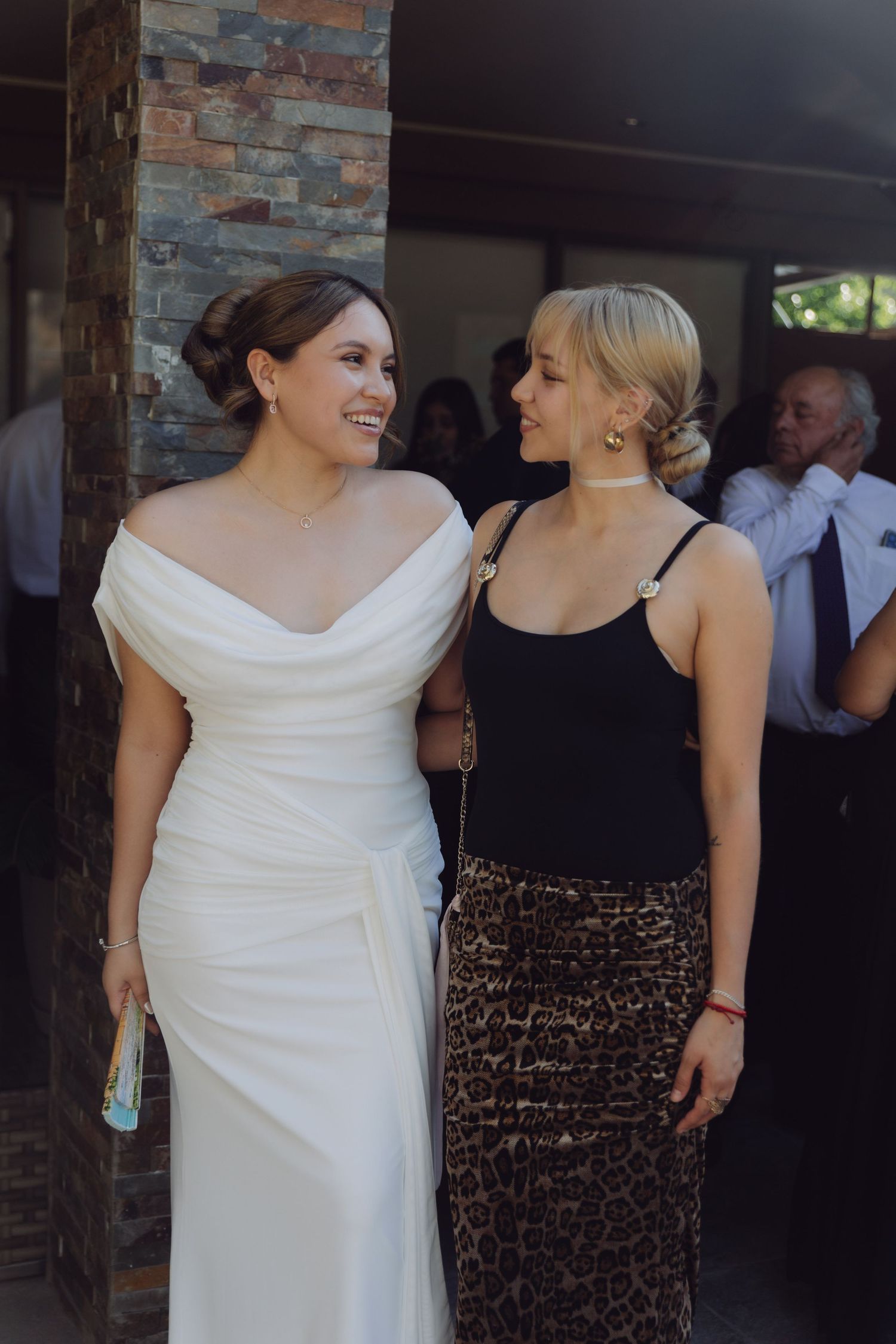 Bride in white off-shoulder gown smiles with friend in black top and leopard skirt at wedding reception.