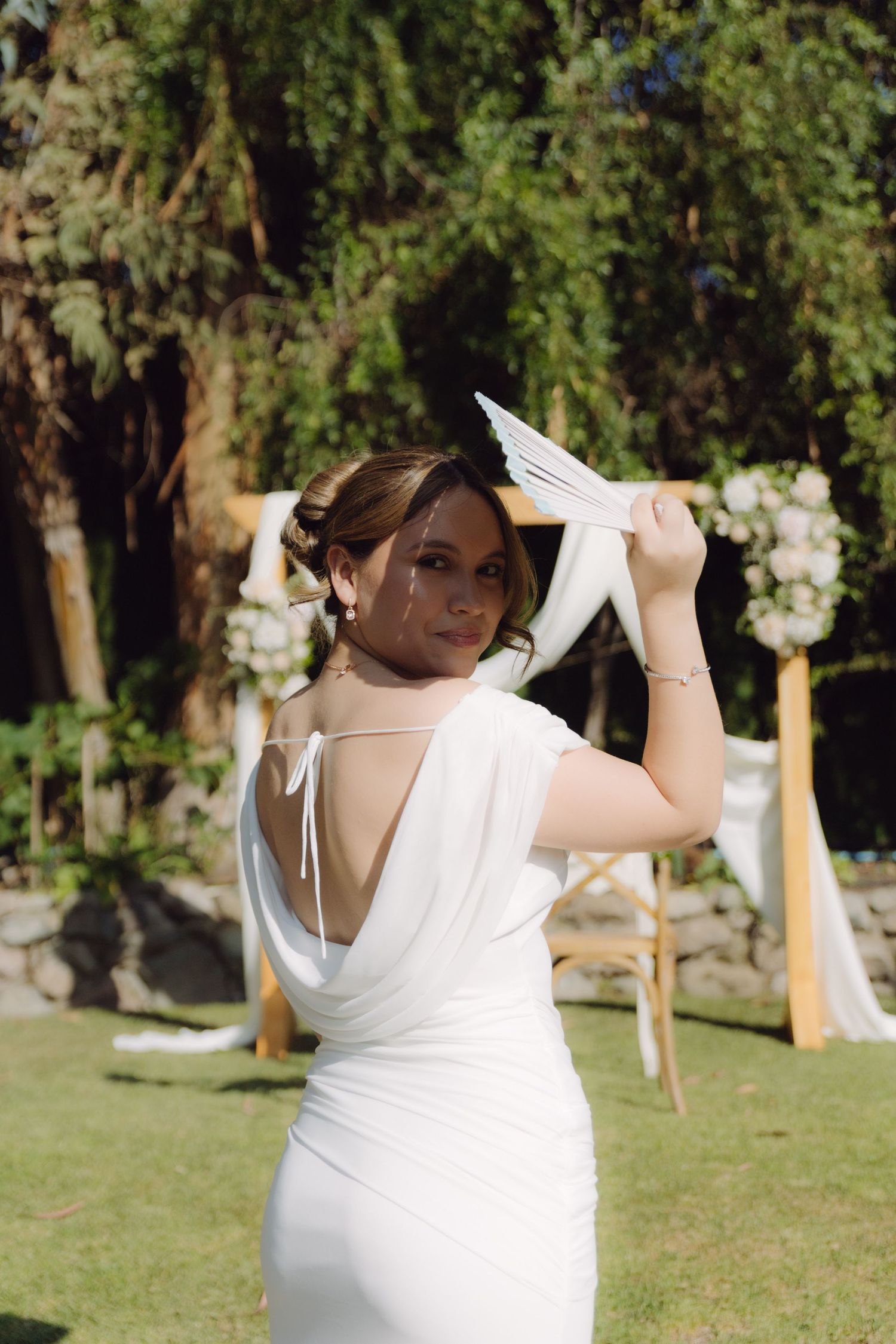 Bride in backless white wedding gown posing with white fan near floral ceremony arch.