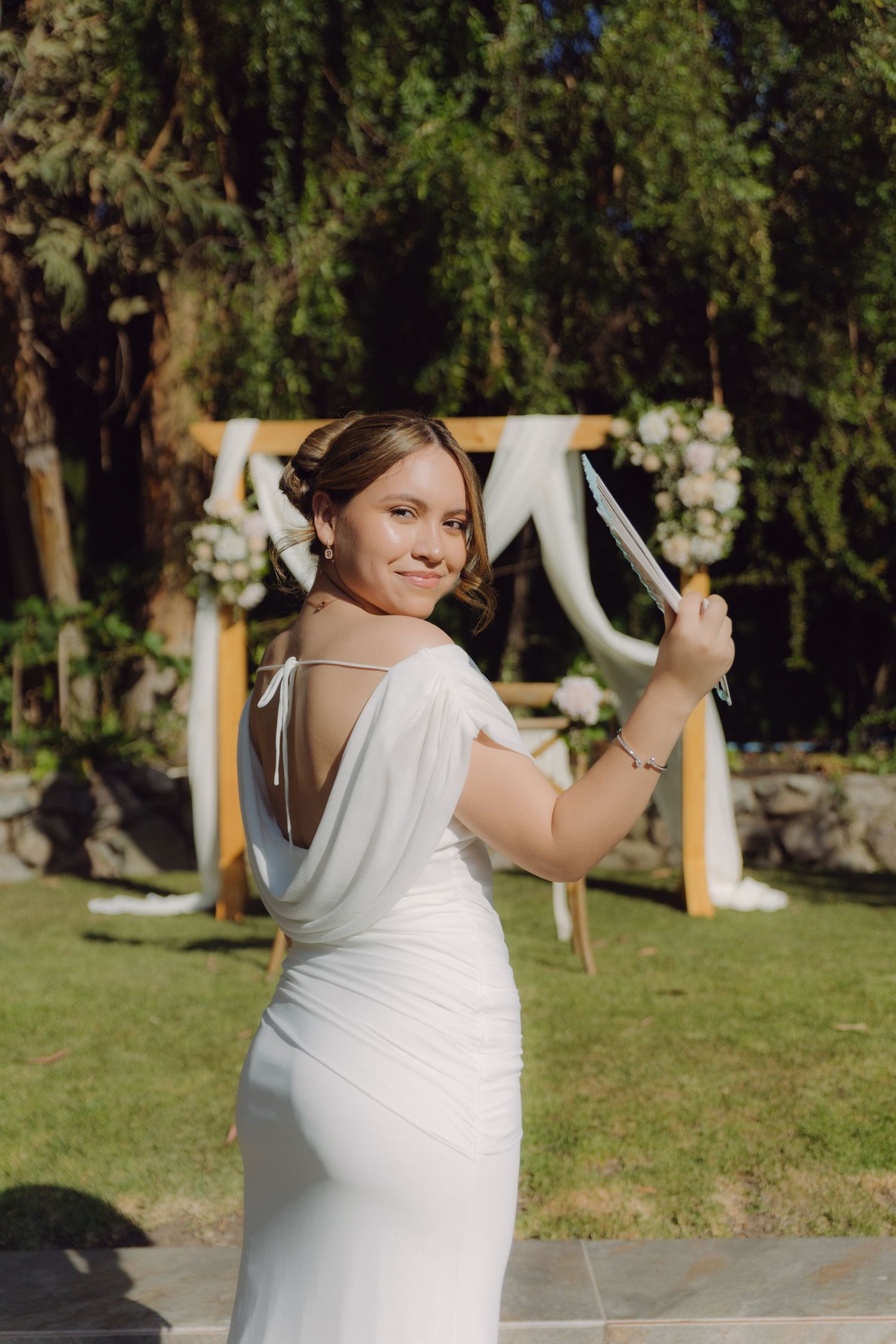 Bride in backless white gown holding fan near floral arch at outdoor wedding ceremony.