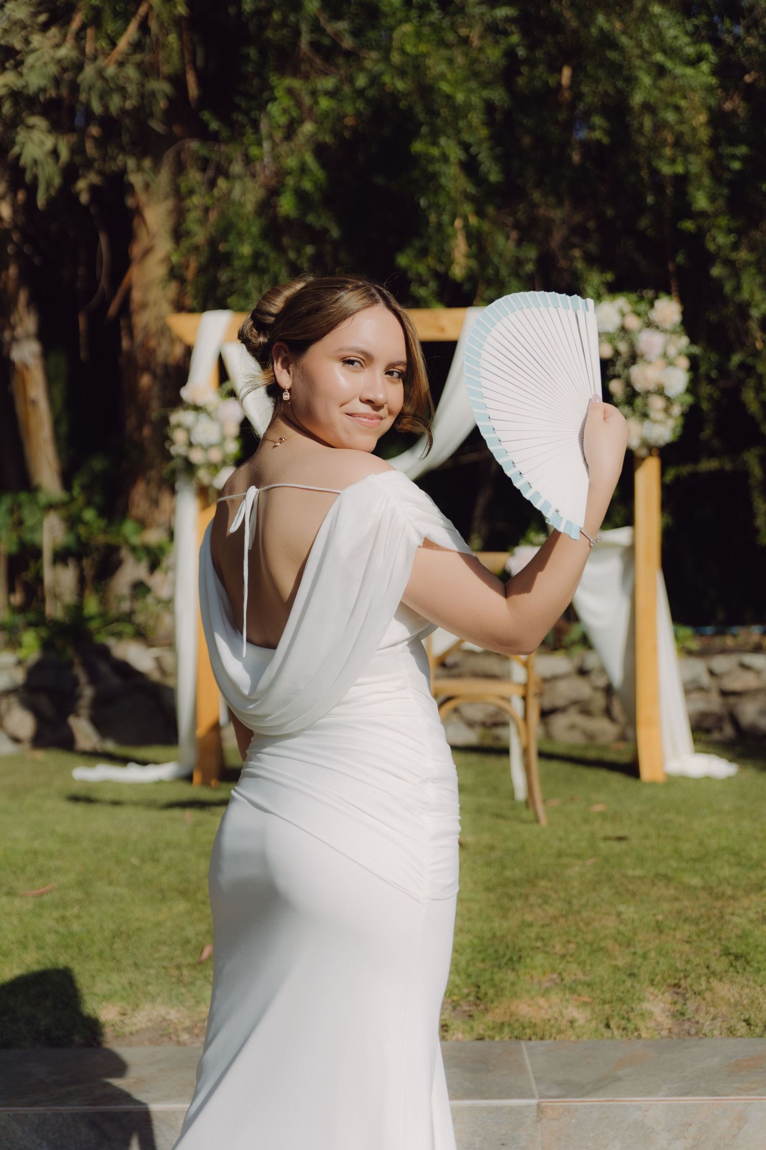 Bride in fitted white backless dress poses with hand fan near a wooden floral arch at outdoor wedding.
