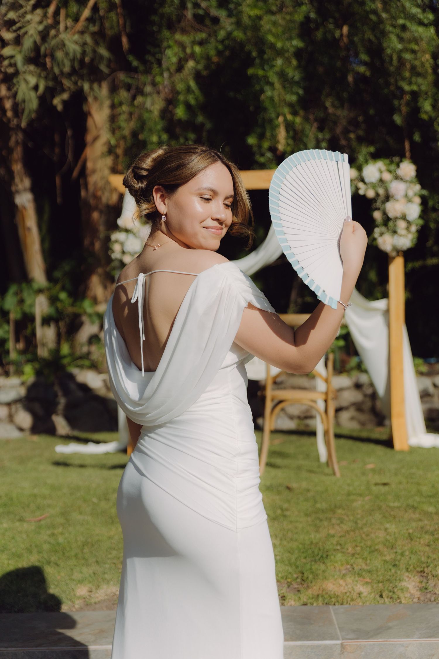 Elegant bride in backless white dress posing with white hand fan at outdoor wedding.