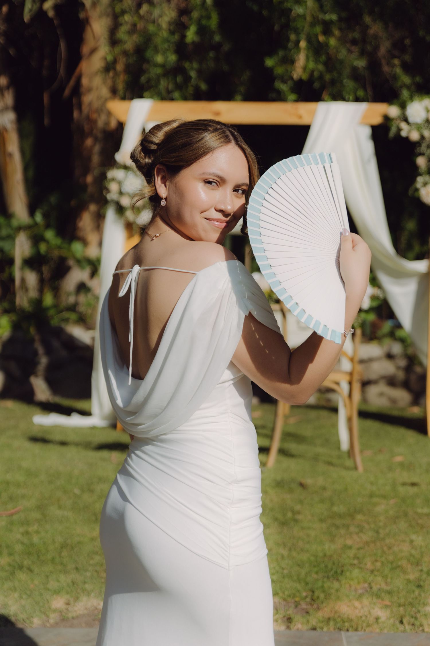 Elegant bride in backless white gown holds a decorative fan outdoors near a floral wedding arch.