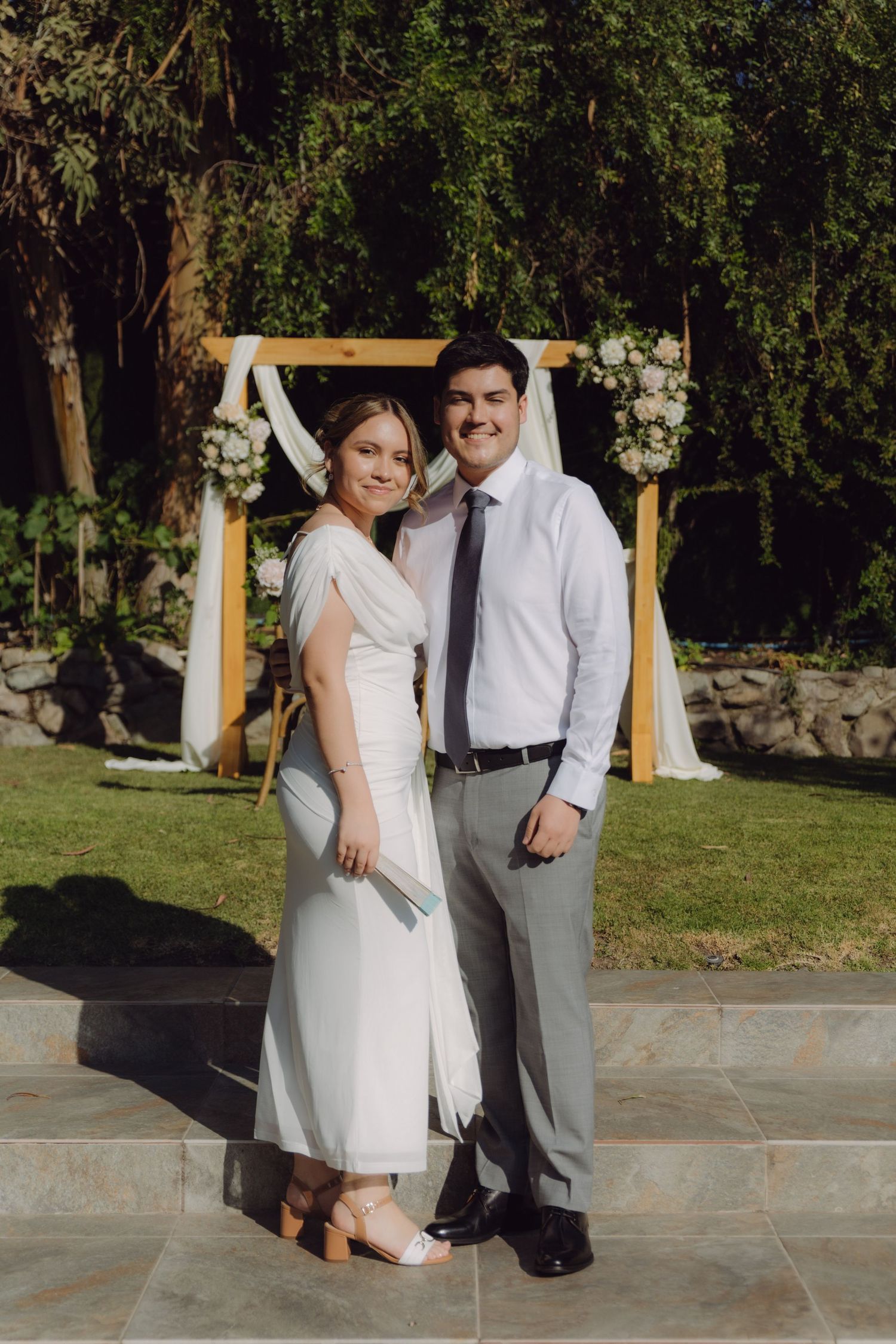 Smiling bride and groom pose together in front of a wooden floral arch at an outdoor wedding ceremony.