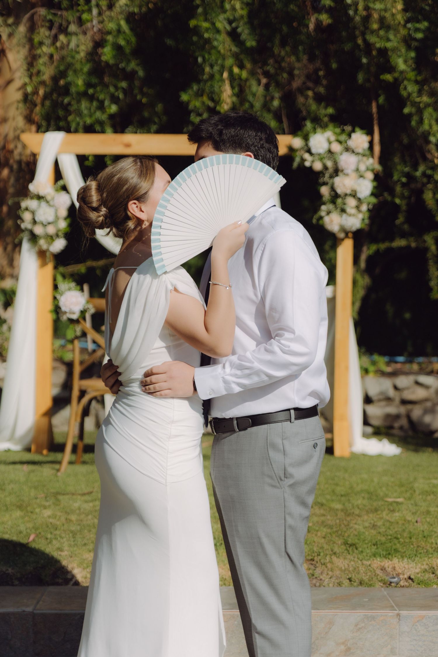 Bride and groom share a kiss hidden behind a white hand fan at their outdoor garden wedding.