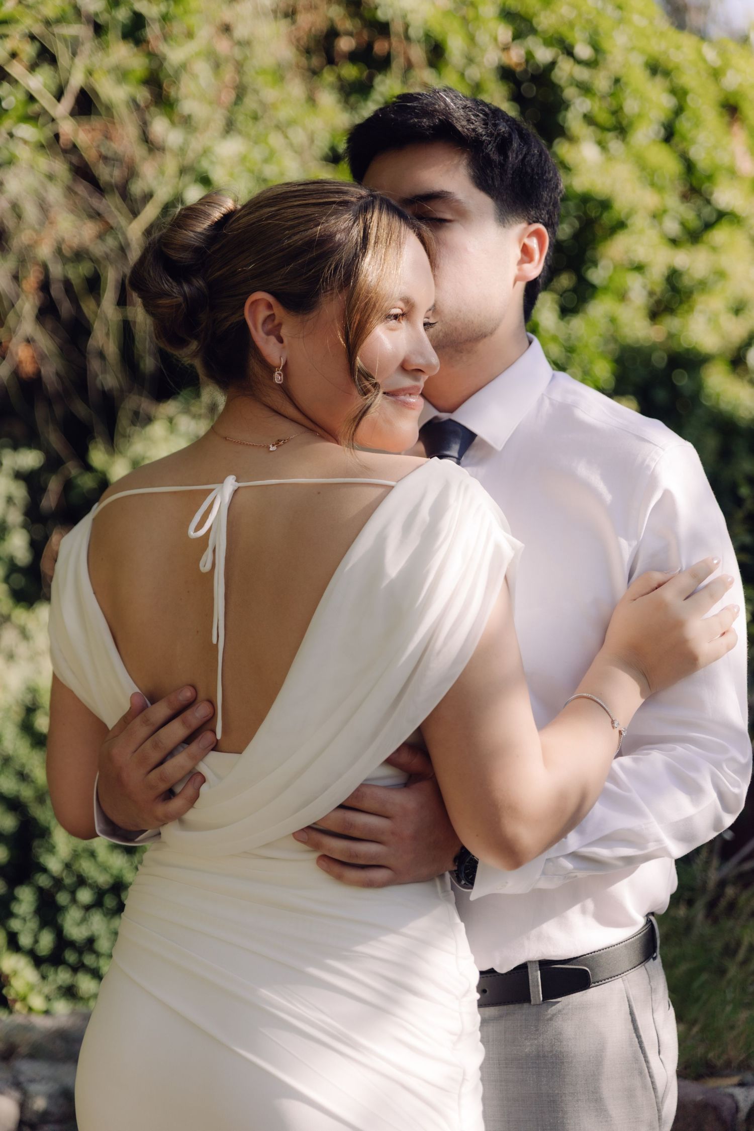 Groom tenderly embraces bride from behind in a backless white gown in a lush garden setting.