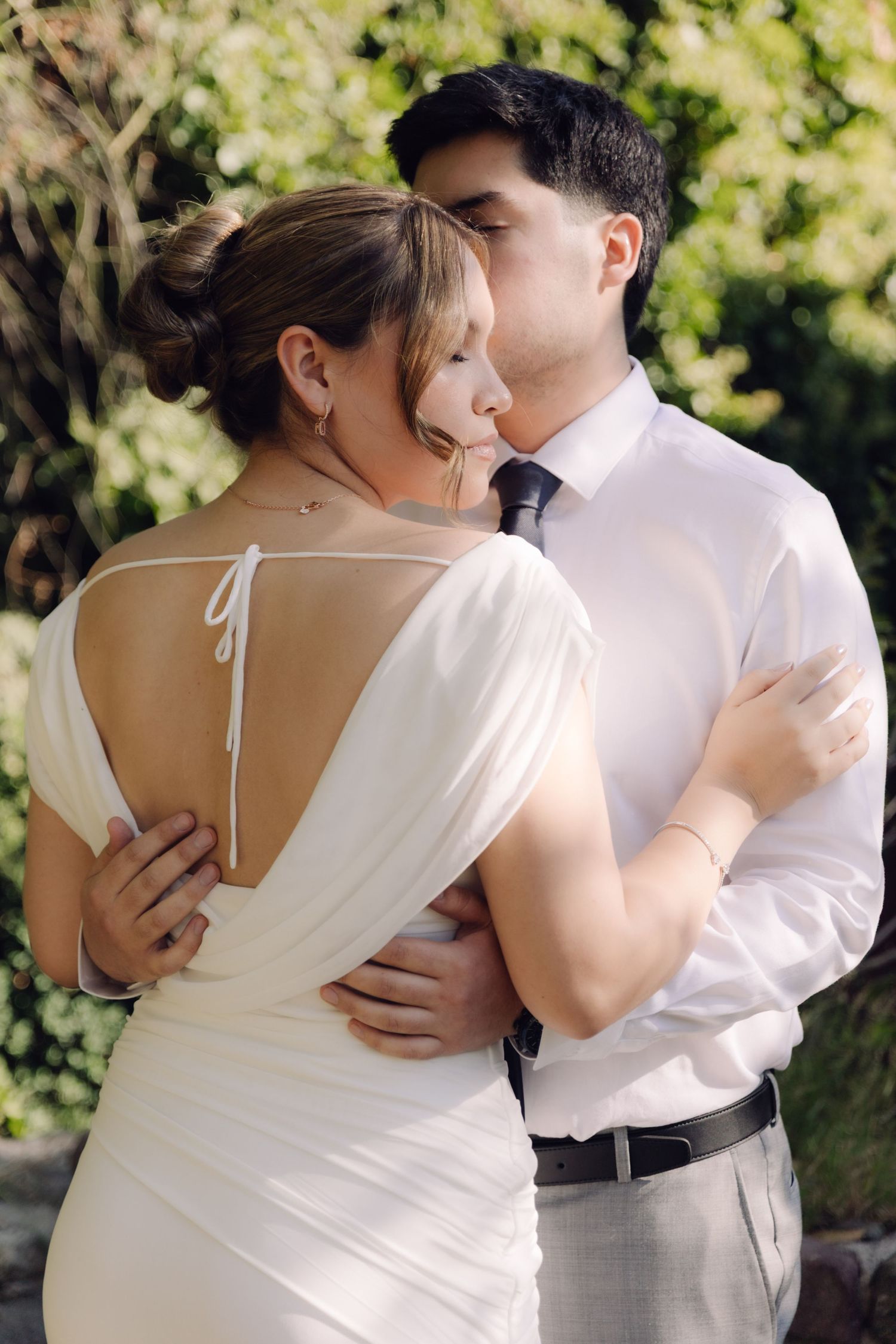 Groom kisses bride on cheek as they embrace outdoors, bride wearing an elegant backless white gown.