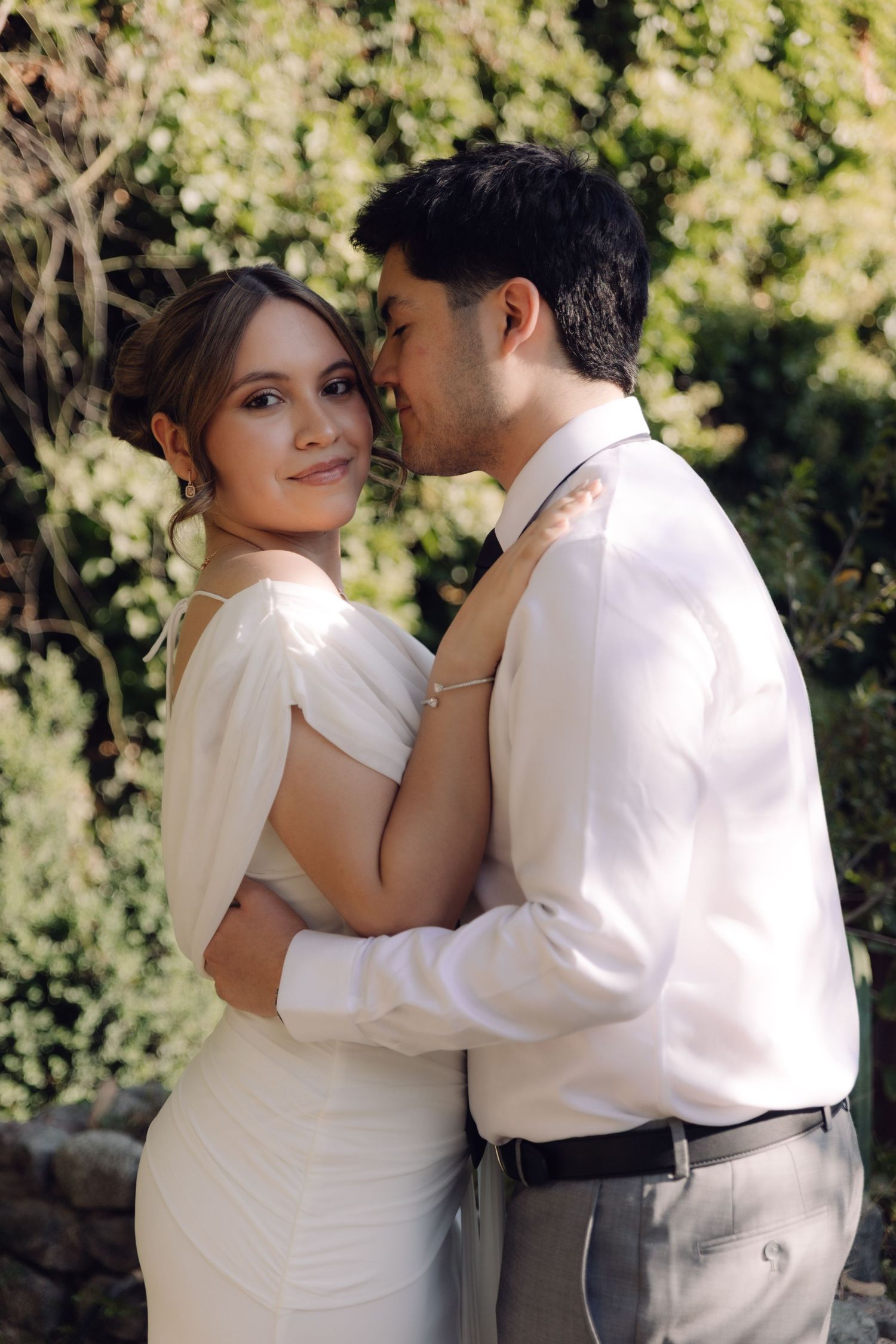 Groom kissing bride on cheek as she smiles at outdoor garden wedding celebration.
