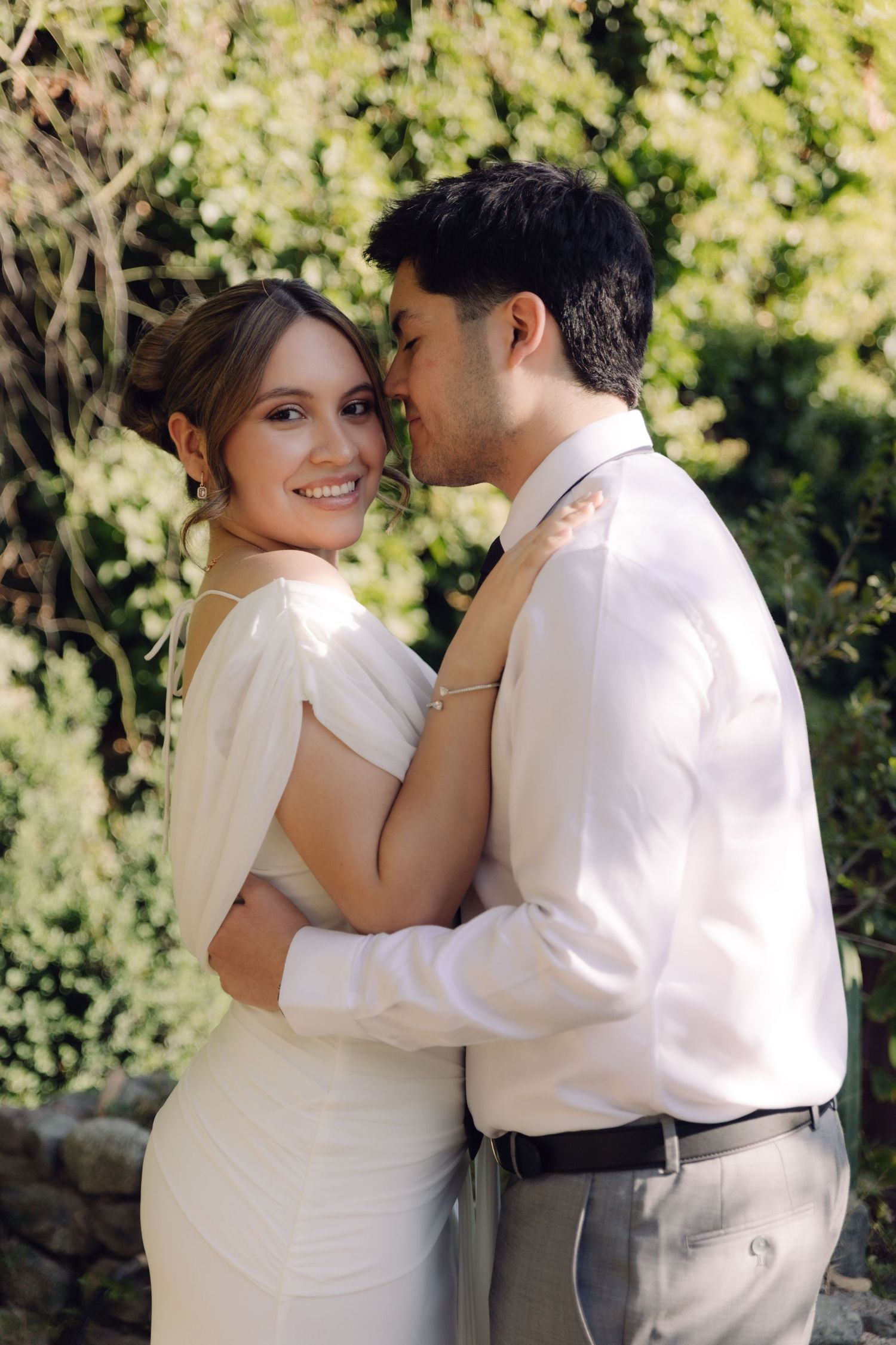 Smiling bride in white dress embraces groom in white shirt outdoors with green foliage background.