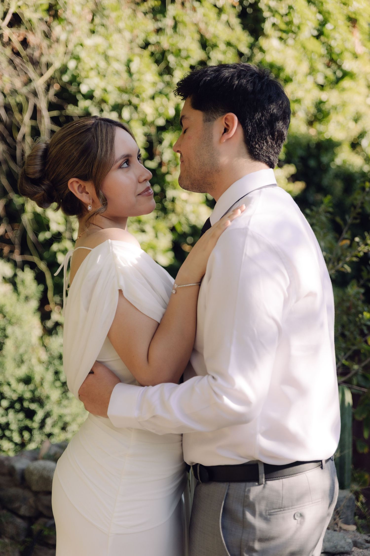 Bride in white gown and groom in white shirt share a romantic gaze while embracing in a garden.