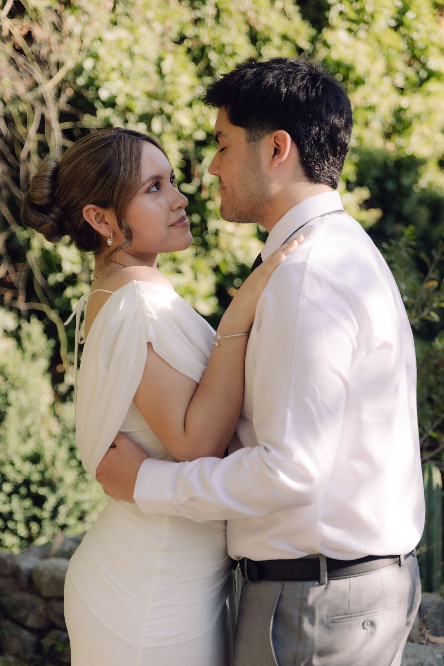 Bride and groom share an intimate gaze while embracing outdoors surrounded by lush greenery.