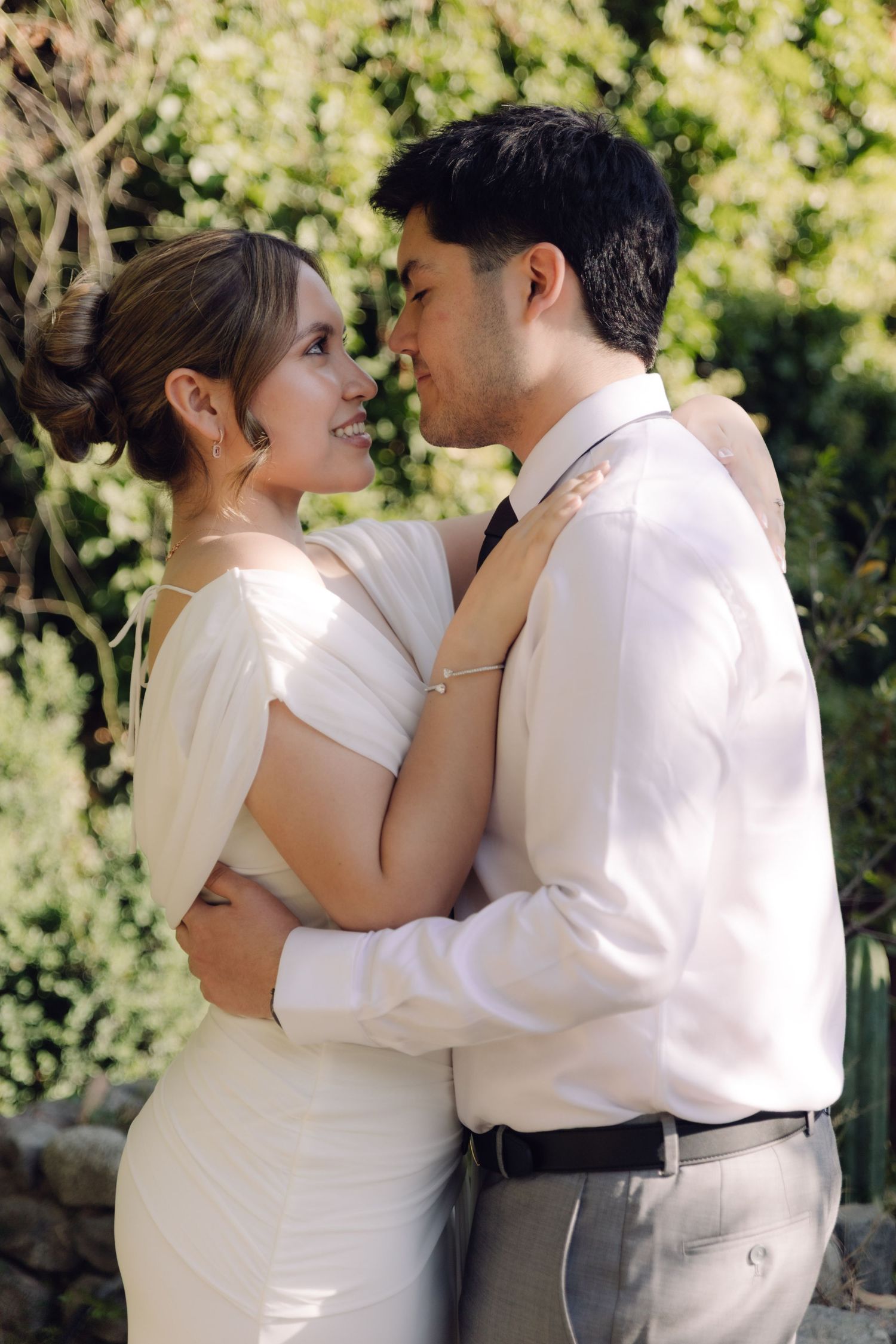 Bride and groom smile at each other in a loving embrace surrounded by lush greenery at their wedding.