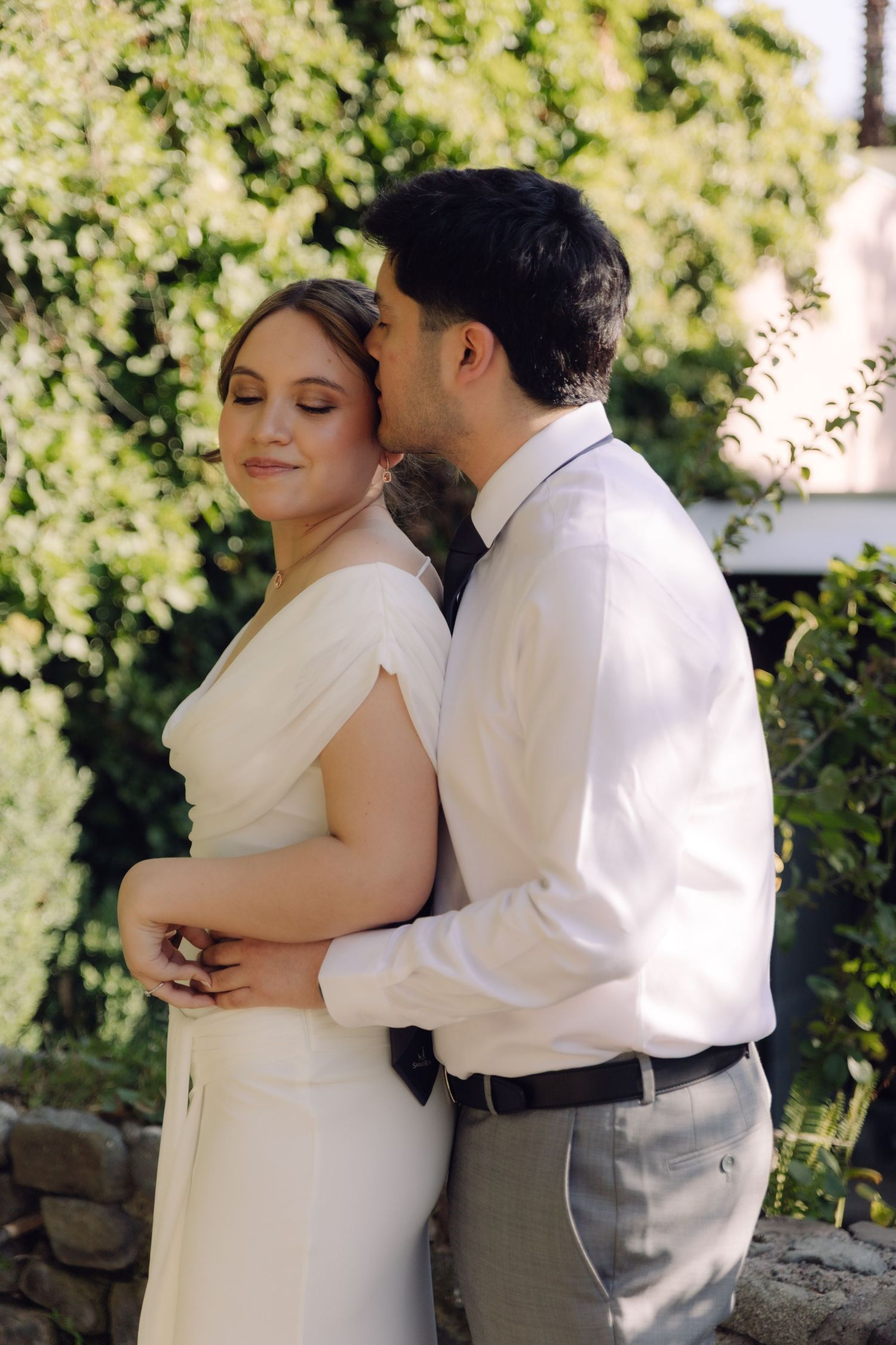 Groom kissing bride on cheek while embracing her outdoors in lush green garden setting.