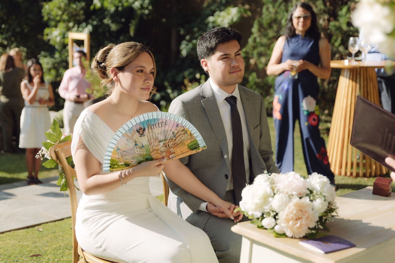 Bride holding decorative fan sits with groom during outdoor wedding ceremony.