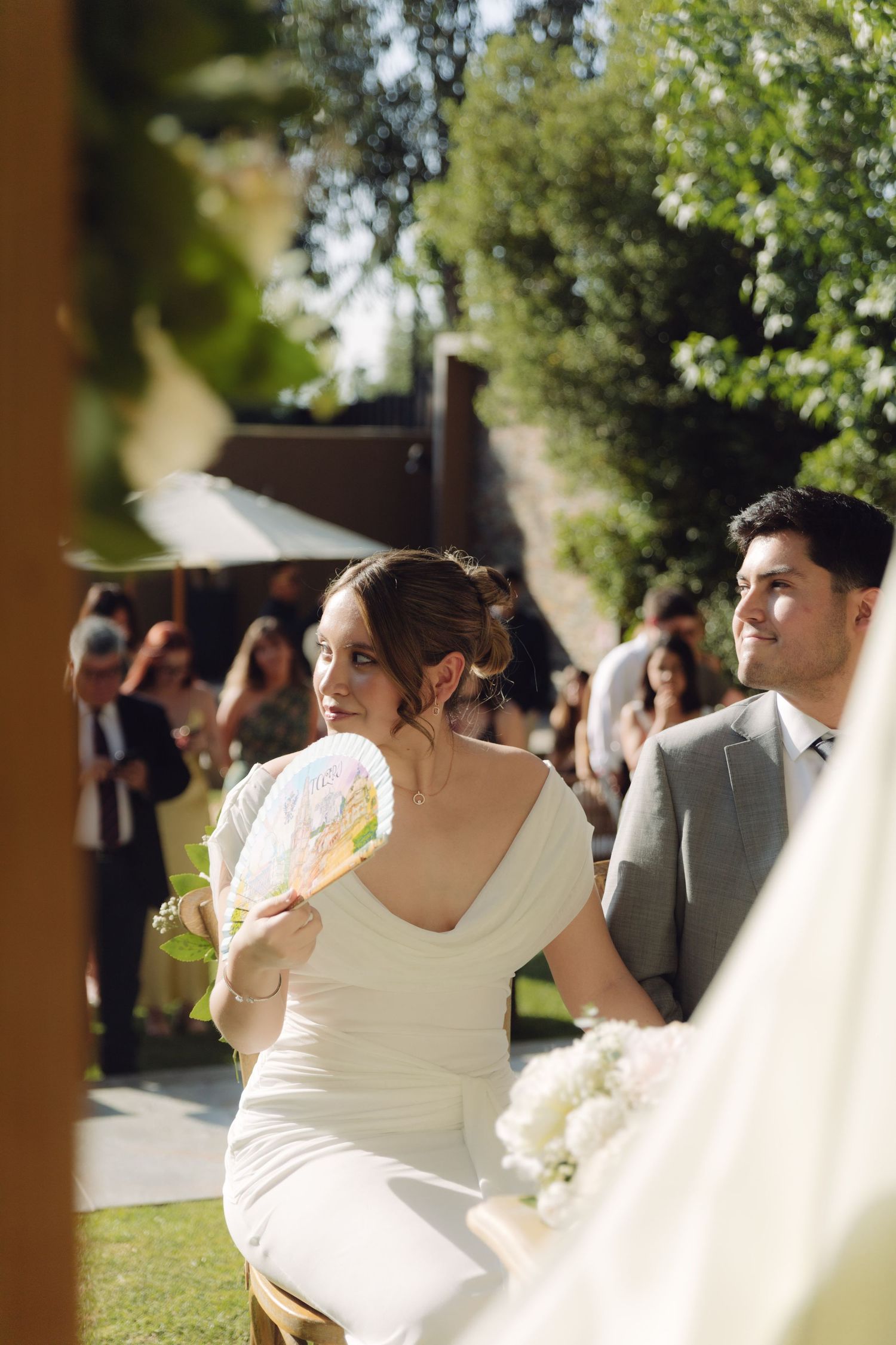 Bride in white dress holds a fan while seated beside groom in gray suit at outdoor wedding.