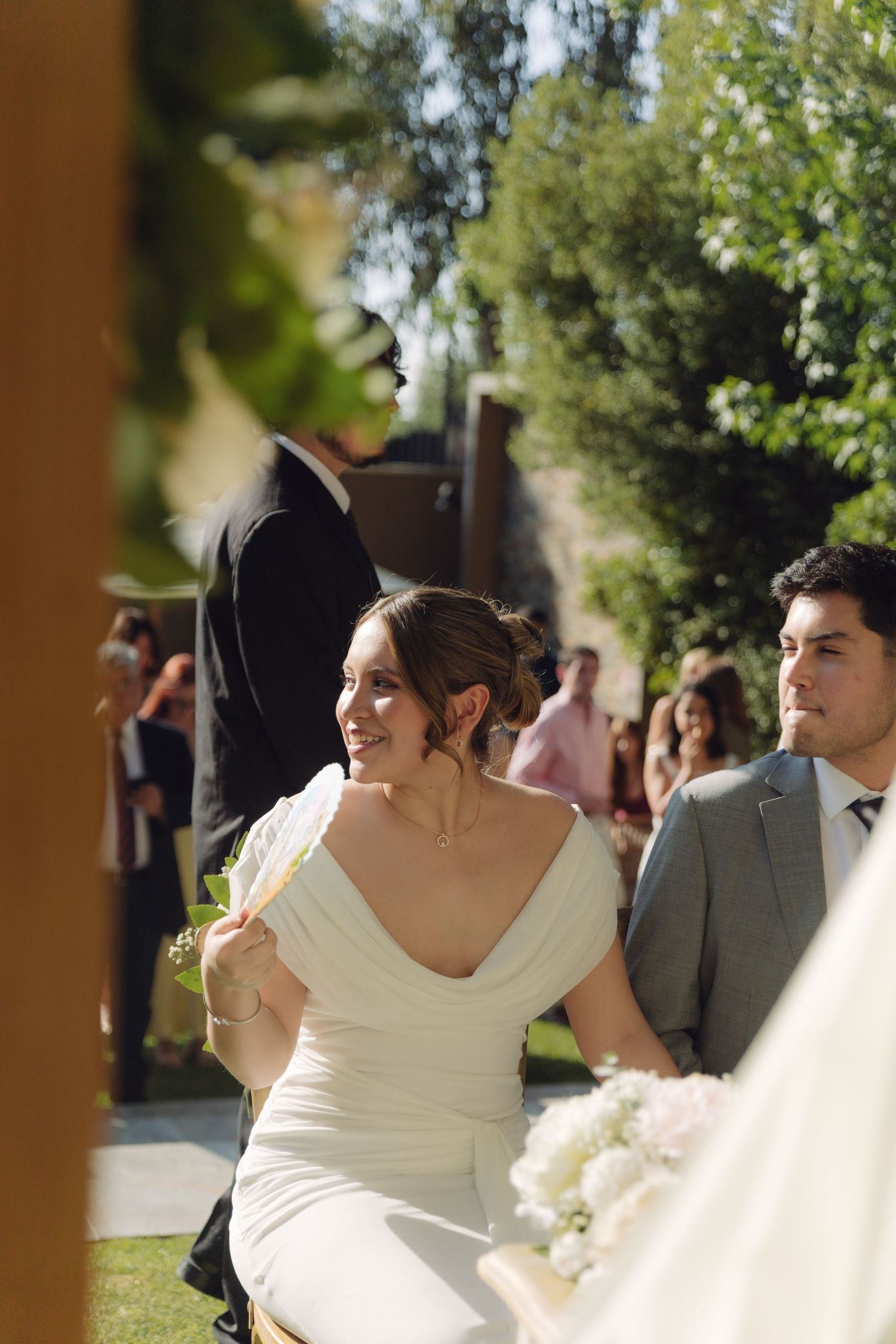 Smiling bride in white gown sits beside groom in gray suit during sunny outdoor wedding ceremony.