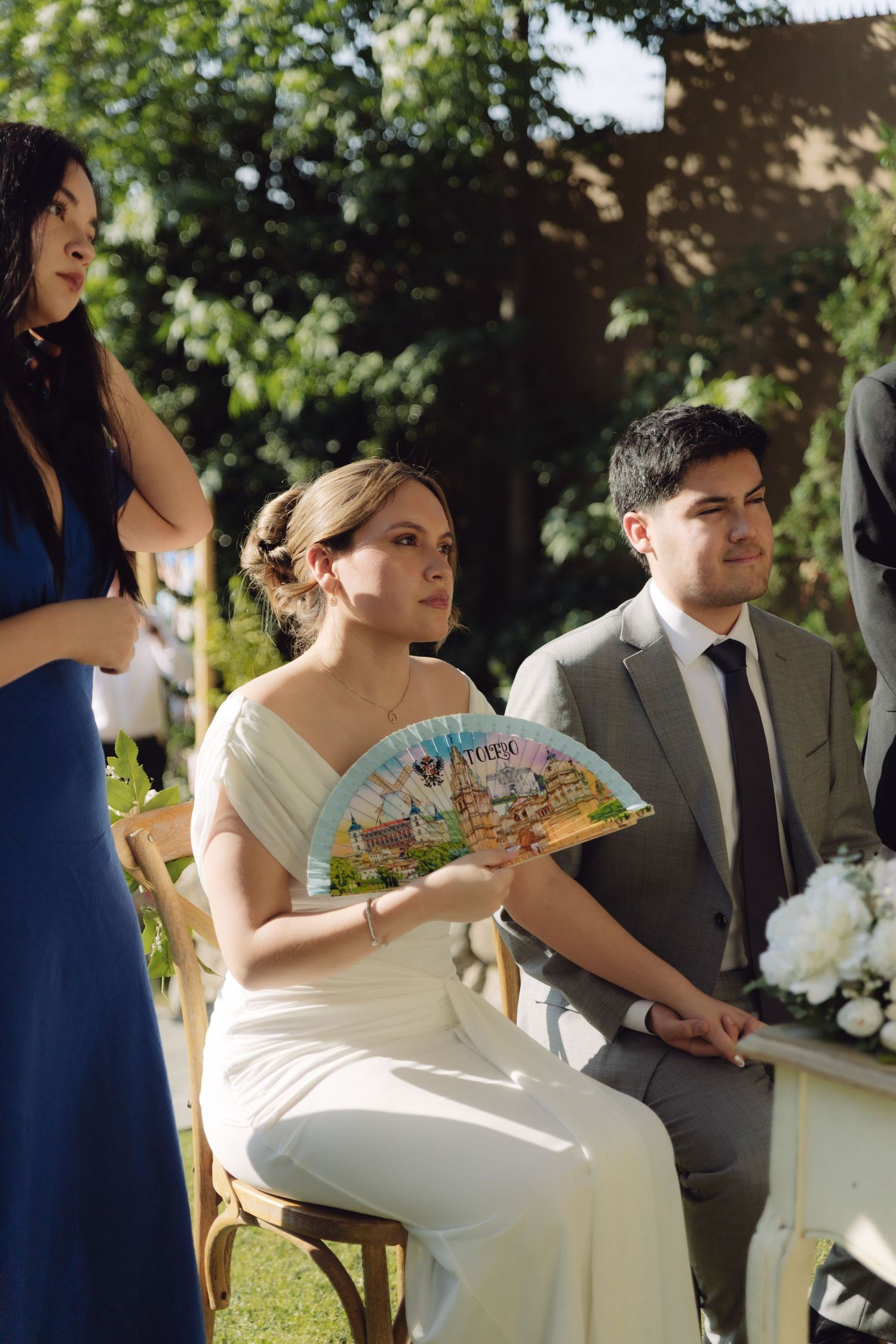Bride fans herself with colorful hand fan beside groom at sunny outdoor wedding.