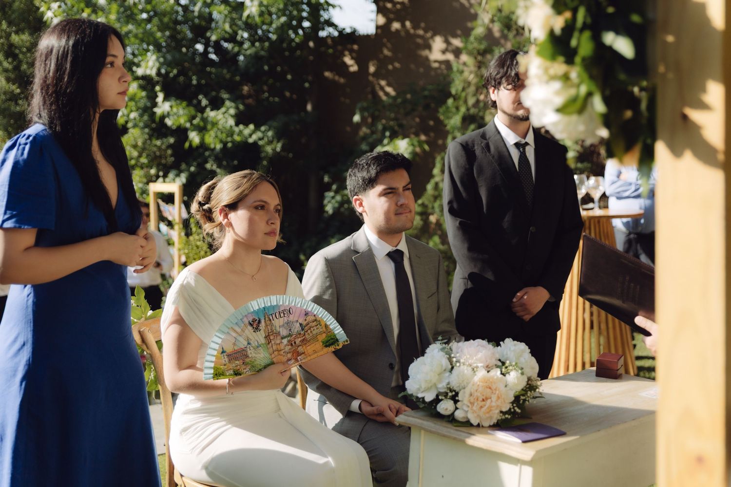 Bride and groom seated at outdoor wedding ceremony with guests and officiant nearby.