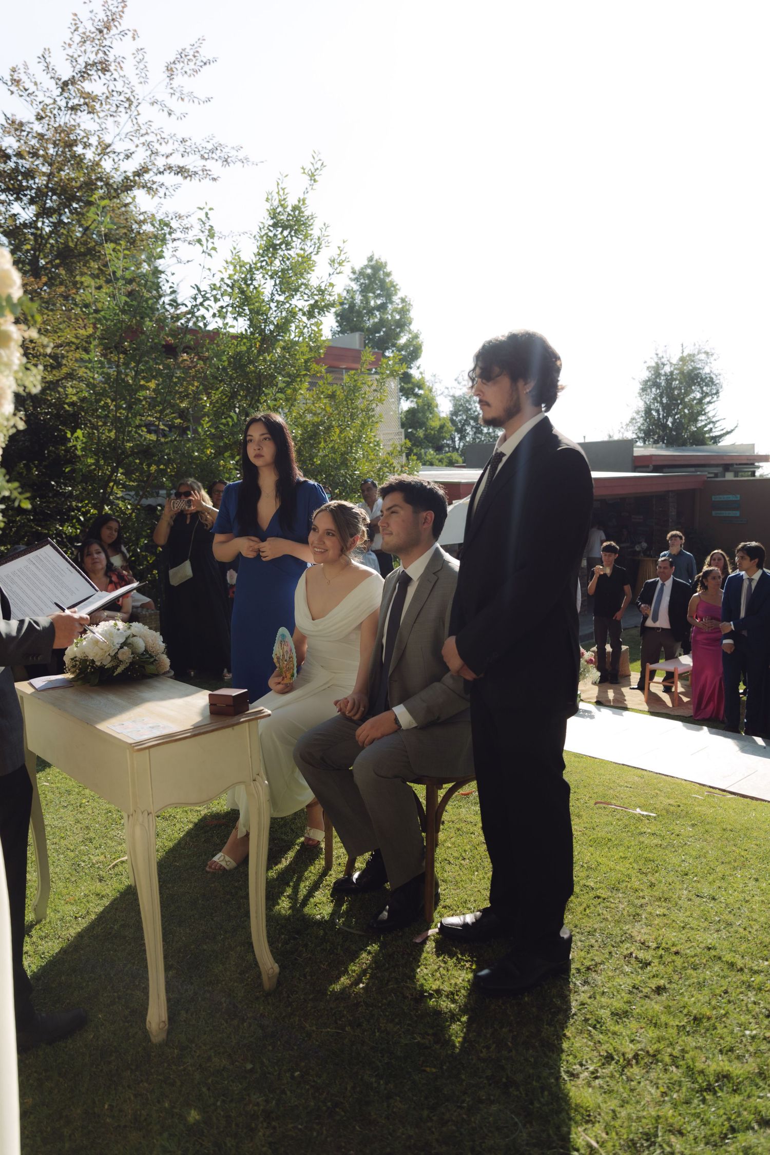 Bride and groom sit at ceremony table during outdoor wedding while guests and officiant stand nearby.