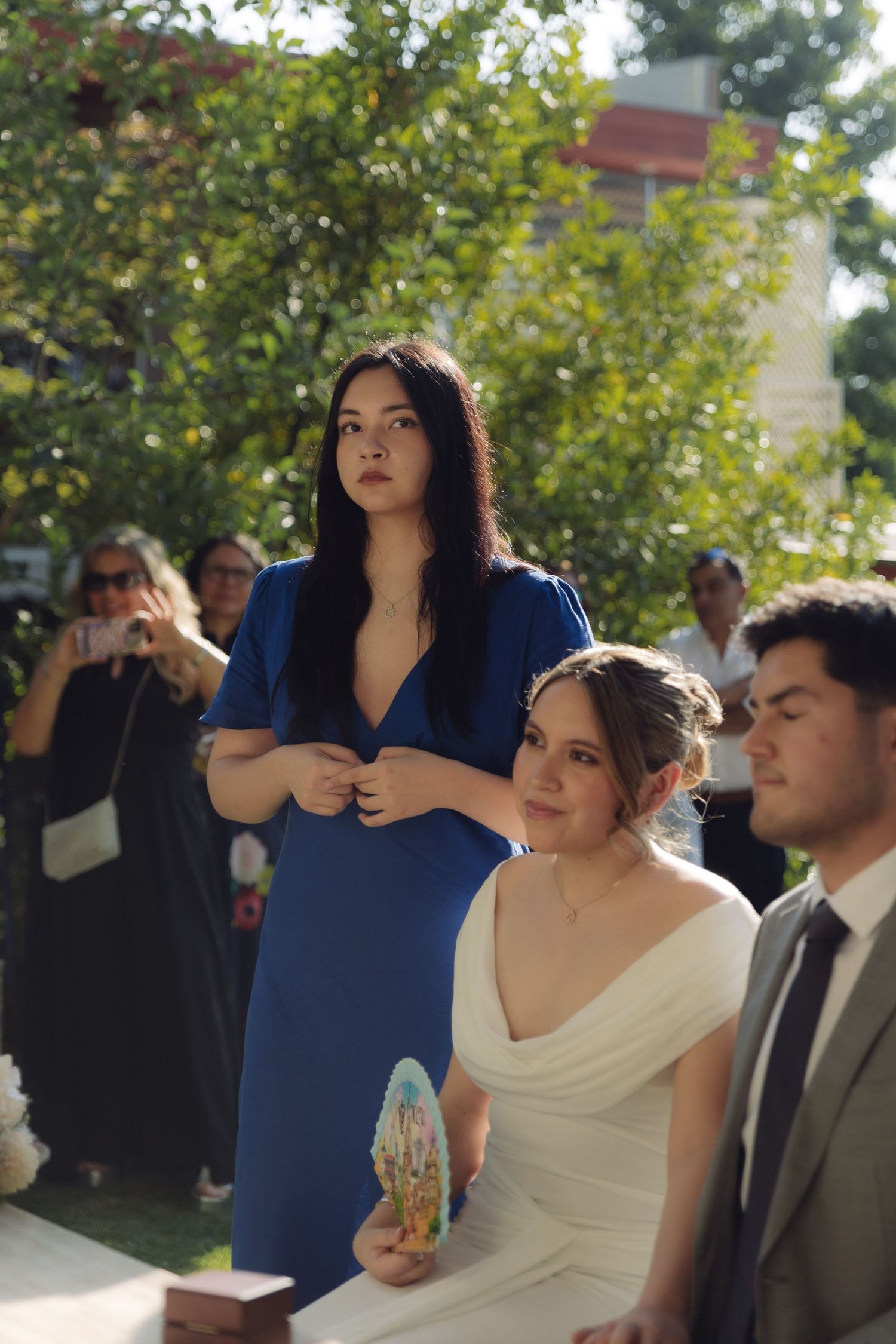 Bride in white dress holds decorative fan beside groom while woman in blue dress stands behind them.