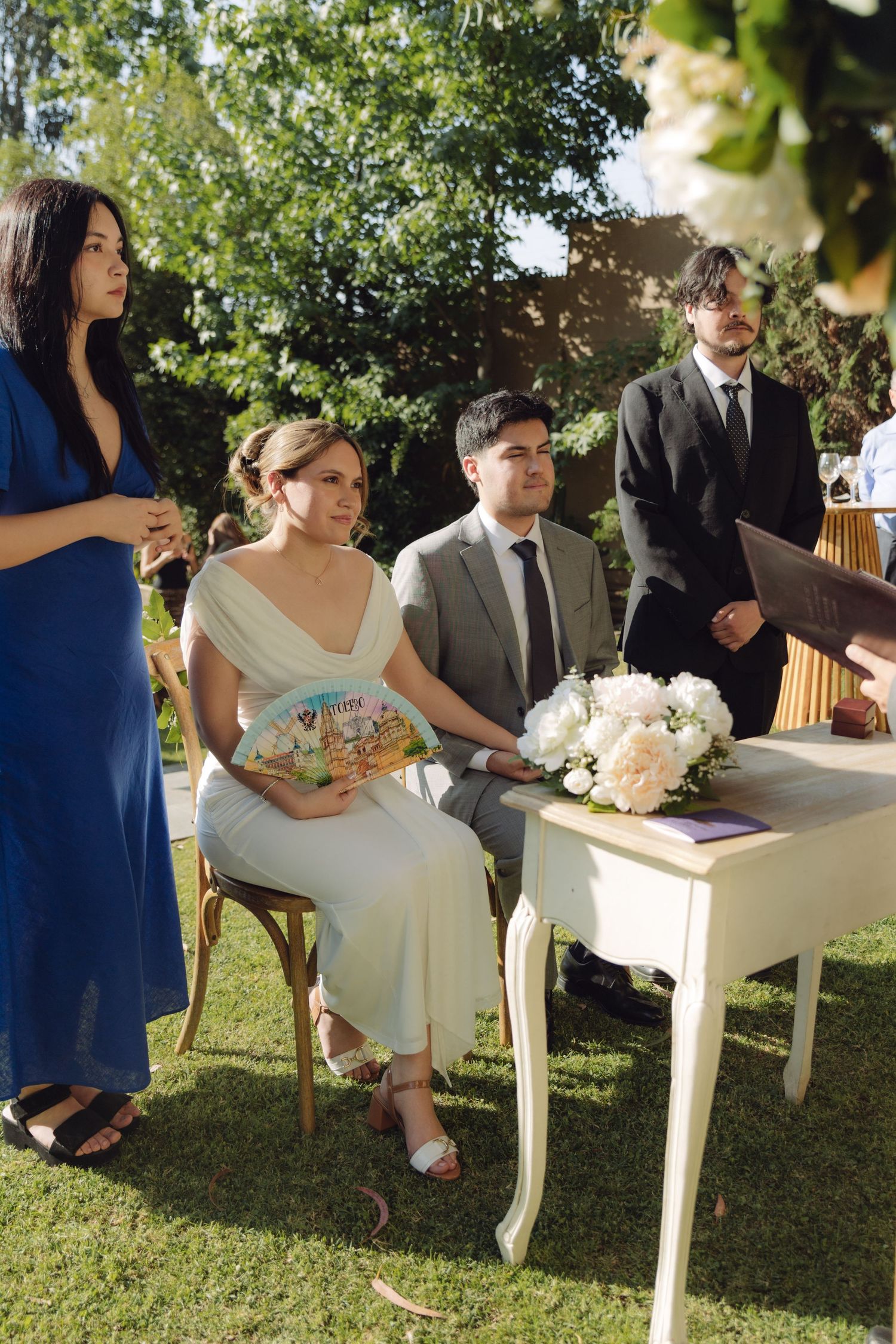 Bride in white gown holds fan beside groom at outdoor wedding ceremony table with white floral bouquet.