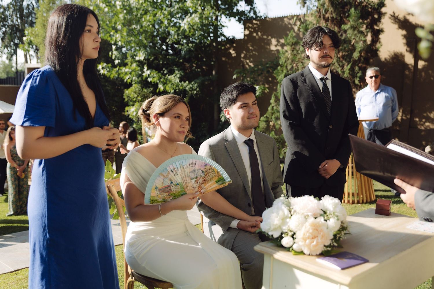 Bride and groom seated at outdoor wedding ceremony with woman in blue dress reading vows.