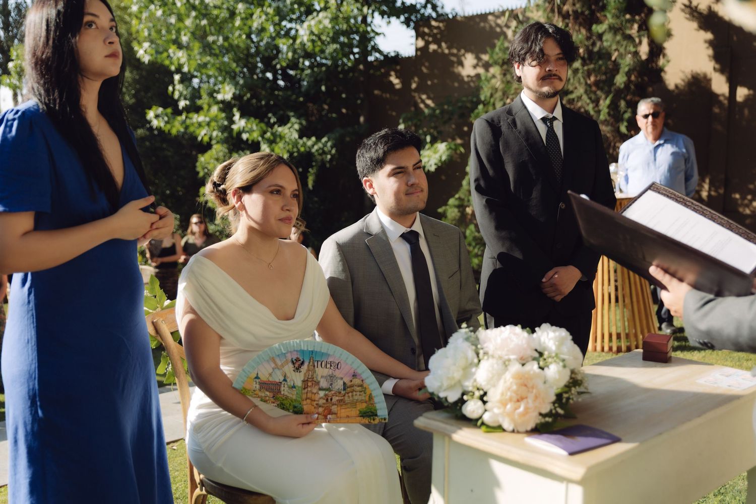 Smiling bride holding colorful book beside groom during sunny outdoor wedding ceremony.