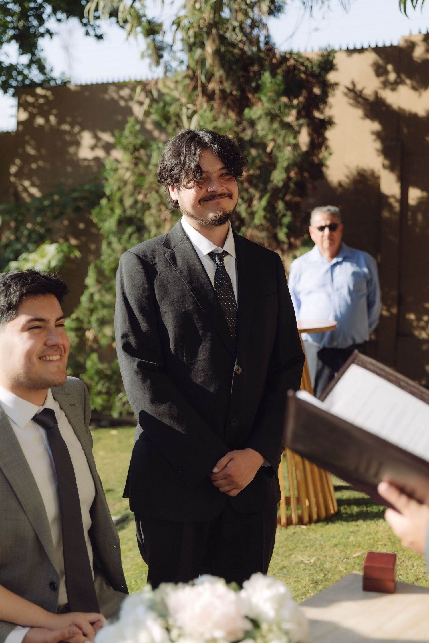 Smiling groomsman in black suit standing at outdoor wedding ceremony altar.