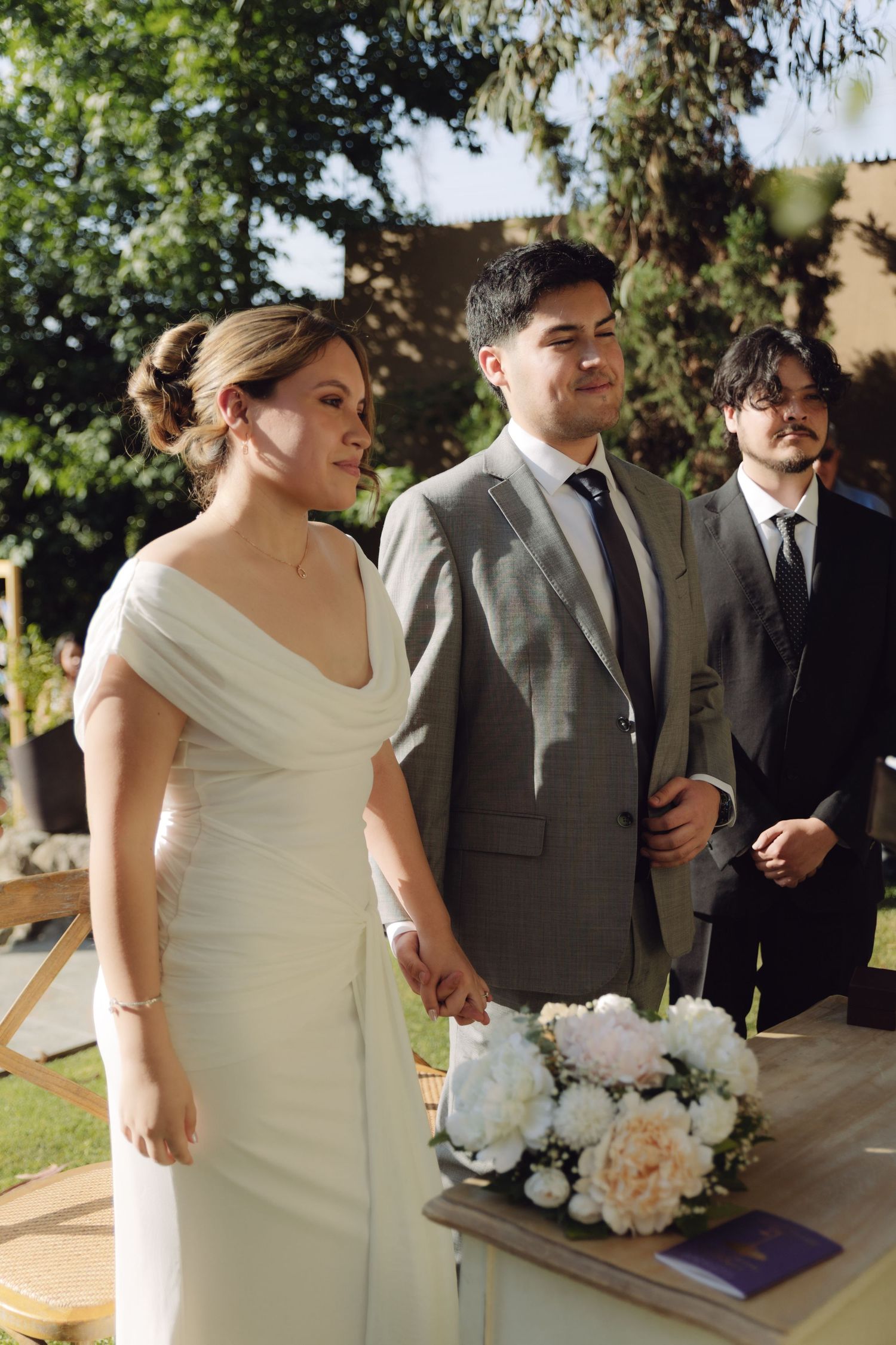 Newlyweds holding hands and smiling beside white floral bouquet at outdoor ceremony.