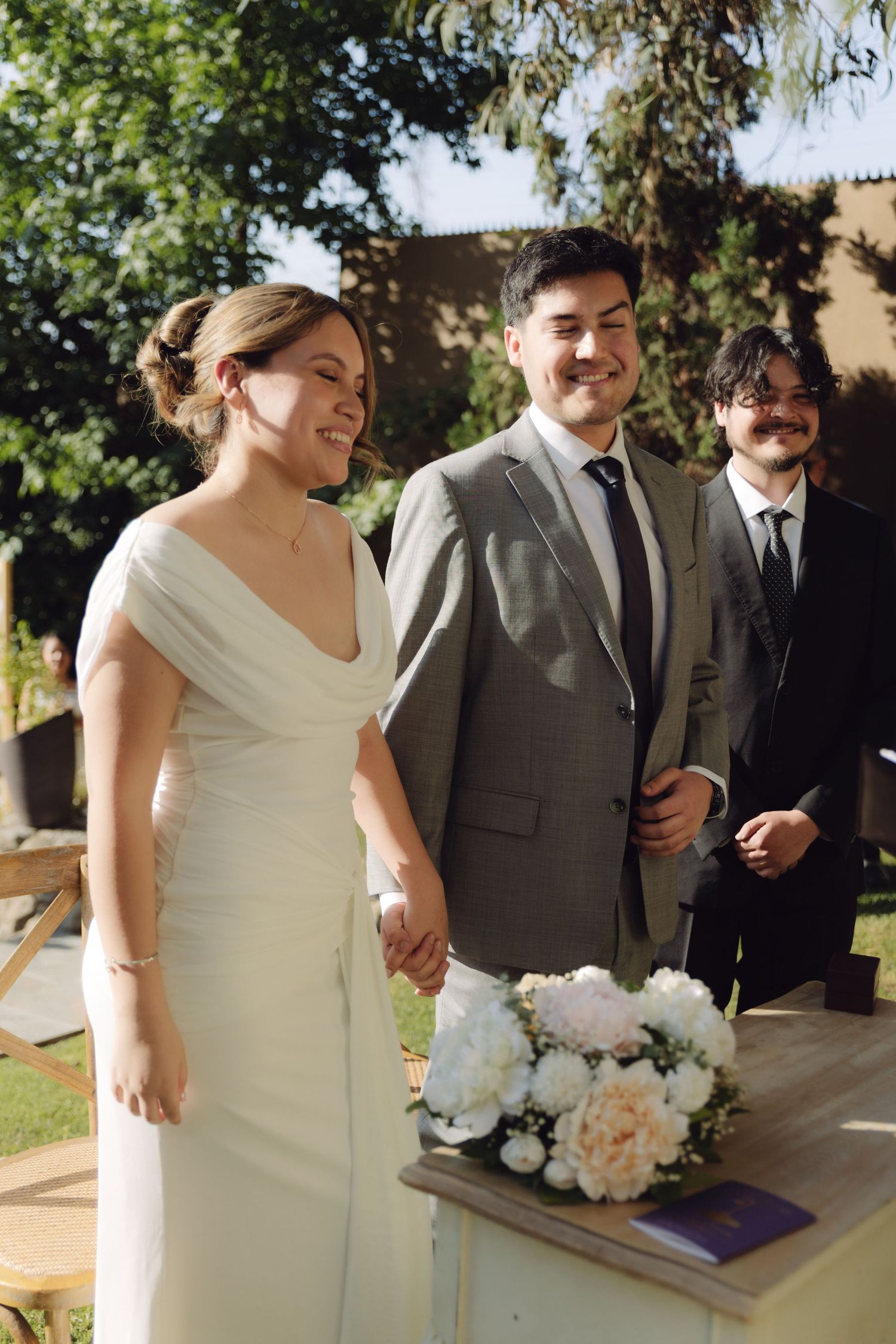 Bride and groom hold hands smiling at outdoor wedding ceremony with floral bouquet on table.